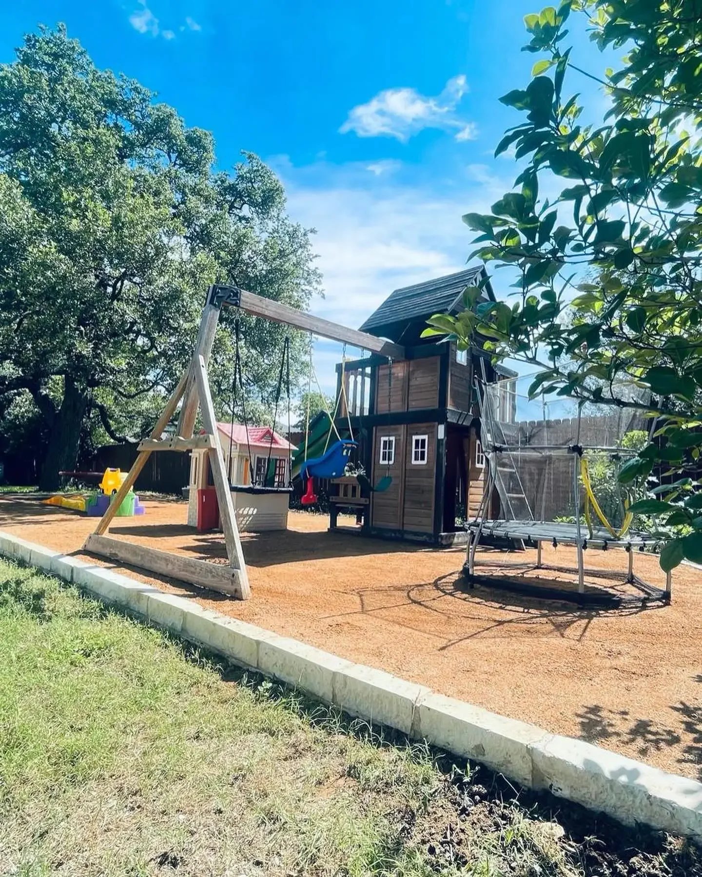 Children's backyard playground with a wooden playhouse, swings, slide, sandbox, and trampoline under a clear blue sky with trees.
