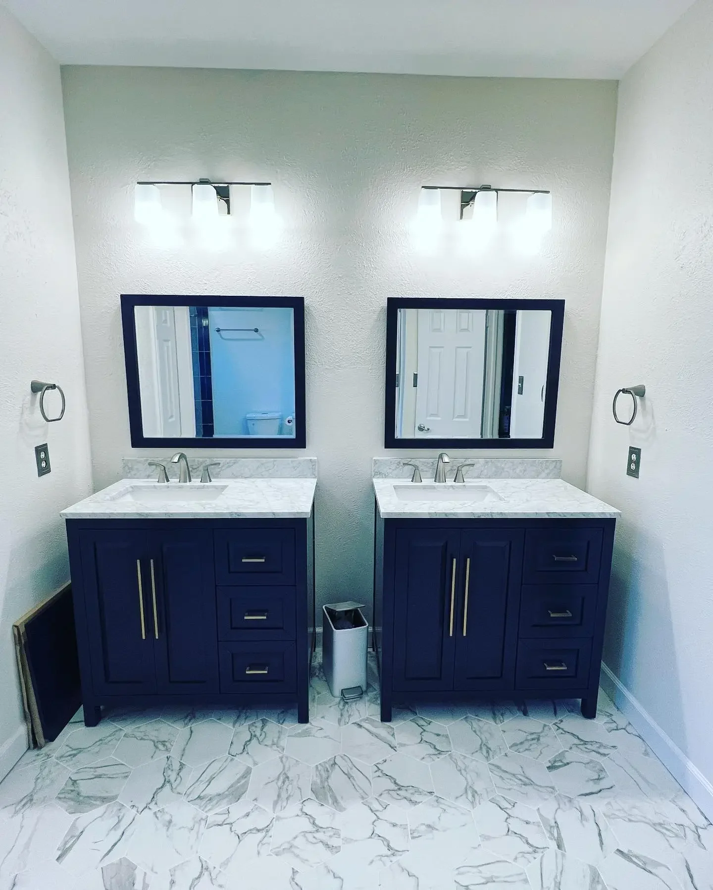 A bathroom with double vanity sinks, two mirrors, and light fixtures. The vanity cabinets are dark blue with gold handles, and the countertops are white marble. There is a small trash bin between the sinks and a framed mirror above each sink. The til
