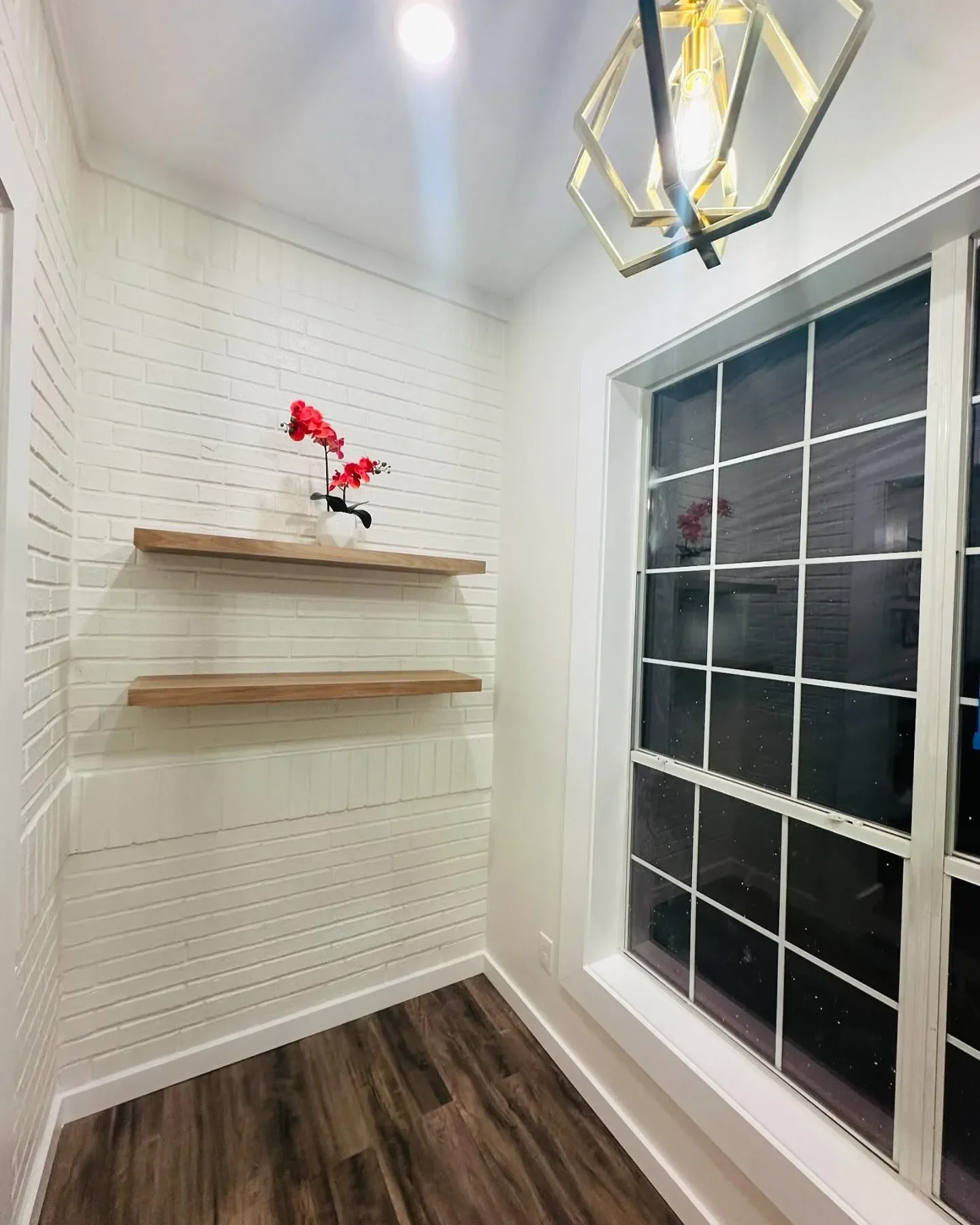Corner of a room with white brick wall, two floating wood shelves, a white vase with red artificial flowers, a large window with white grid frame, and a modern geometric chandelier.