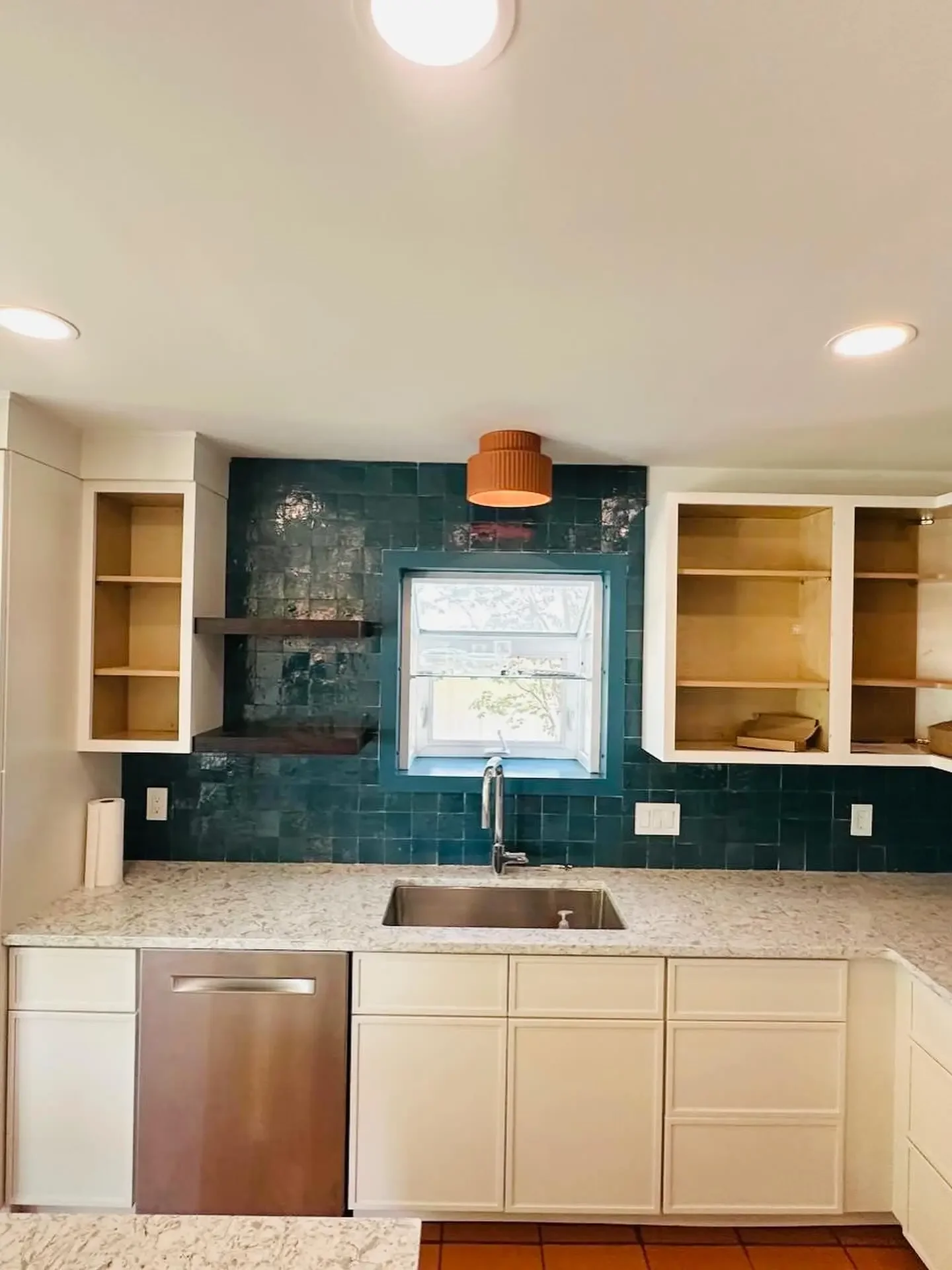 Kitchen with cream cabinets, granite countertop, undermount sink, dark green tile backsplash, open wooden shelves, small window above the sink, ceiling lights, and terracotta floor tiles.
