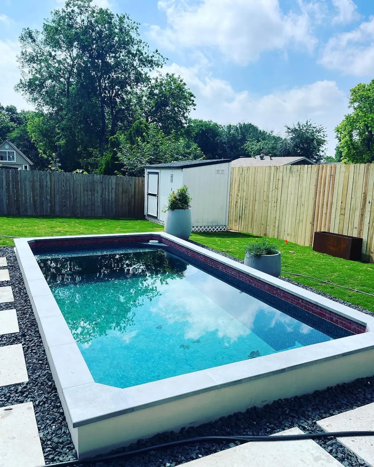 Backyard with a small rectangular swimming pool, wooden fence, green lawn, potted plants, a white storage shed, and trees under a partly cloudy sky.