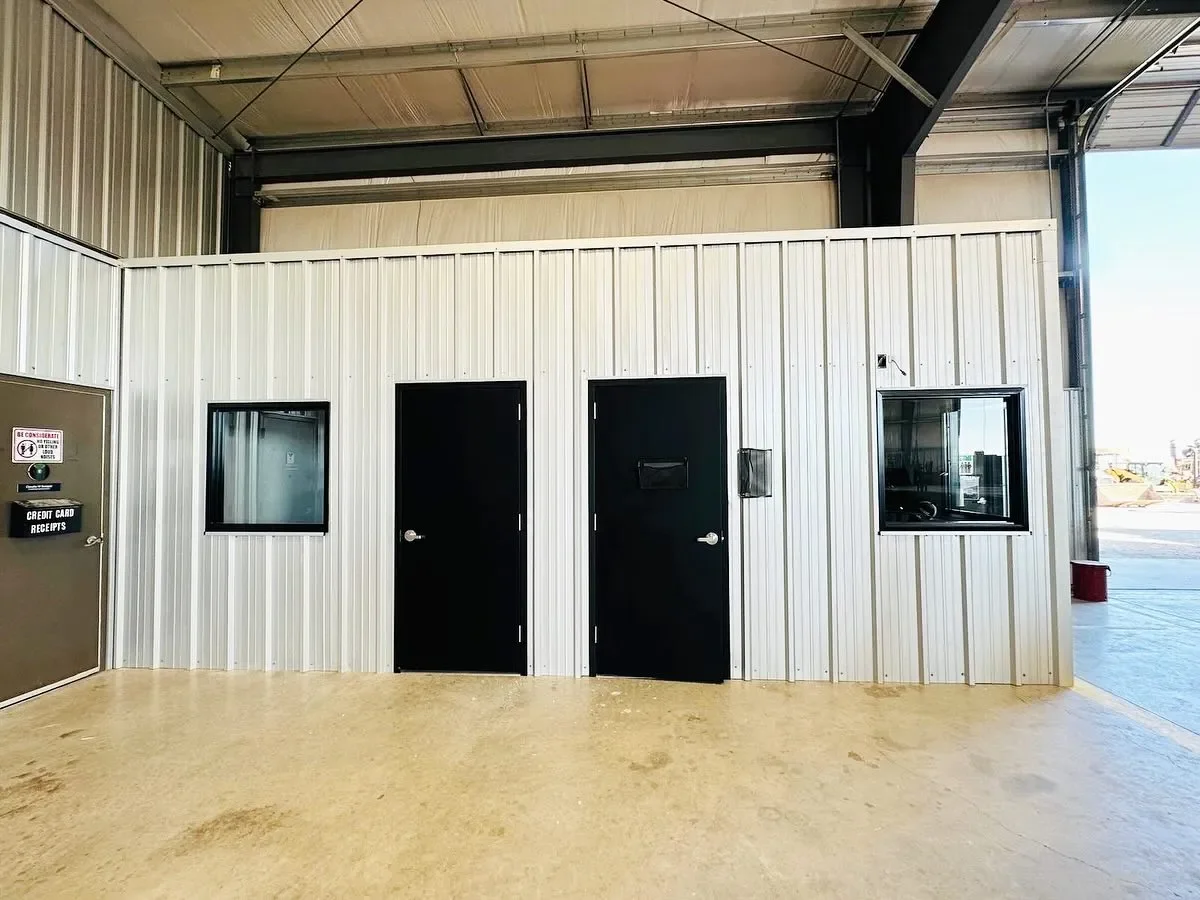 Interior view of a metal building with three black doors and two windows, one on each side of the doors, and a small mail slot on one door. The floor is concrete, and the ceiling is exposed metal beams.