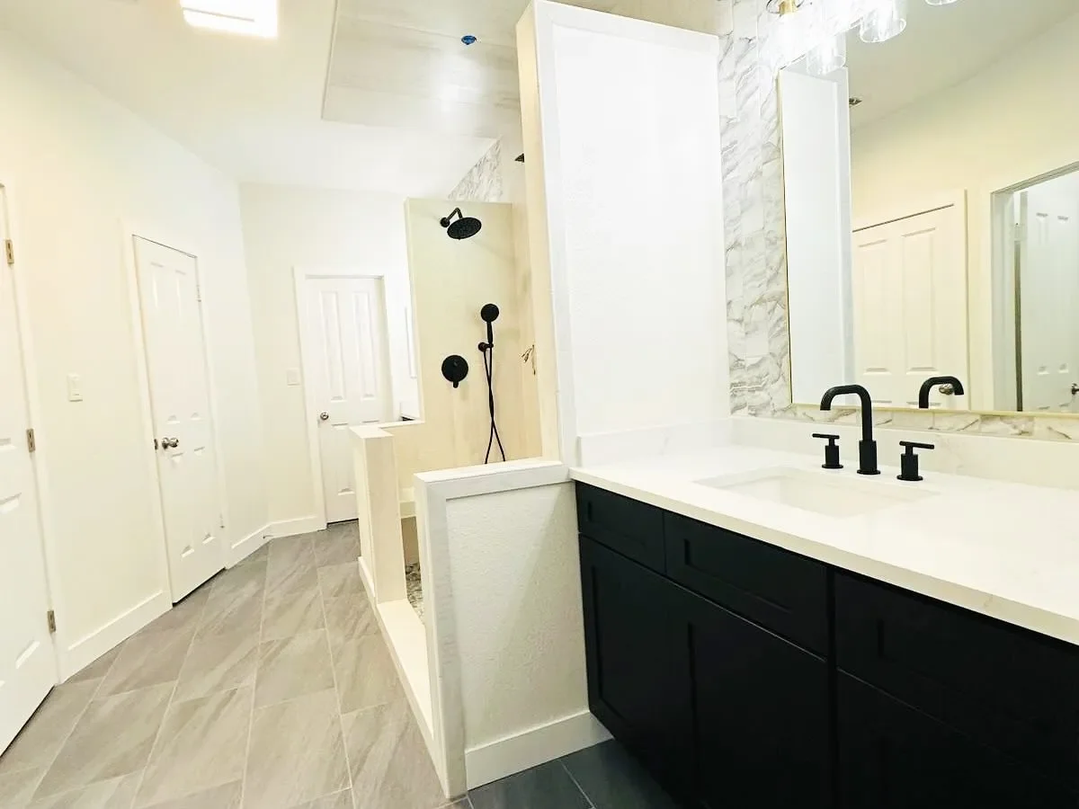 Modern bathroom with a white vanity, black fixtures, a large mirror, a marble backsplash, a walk-in shower with black shower fixtures, and white doors.