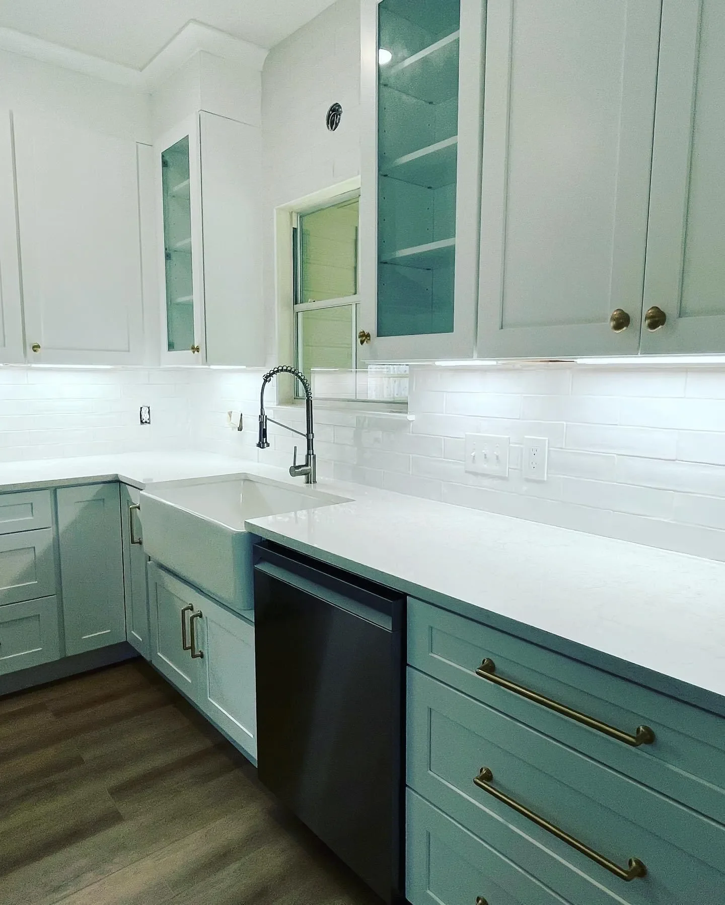A modern kitchen with white cabinets, a white farmhouse sink, and a white countertop. There are brass handles on the cabinets and wood flooring.