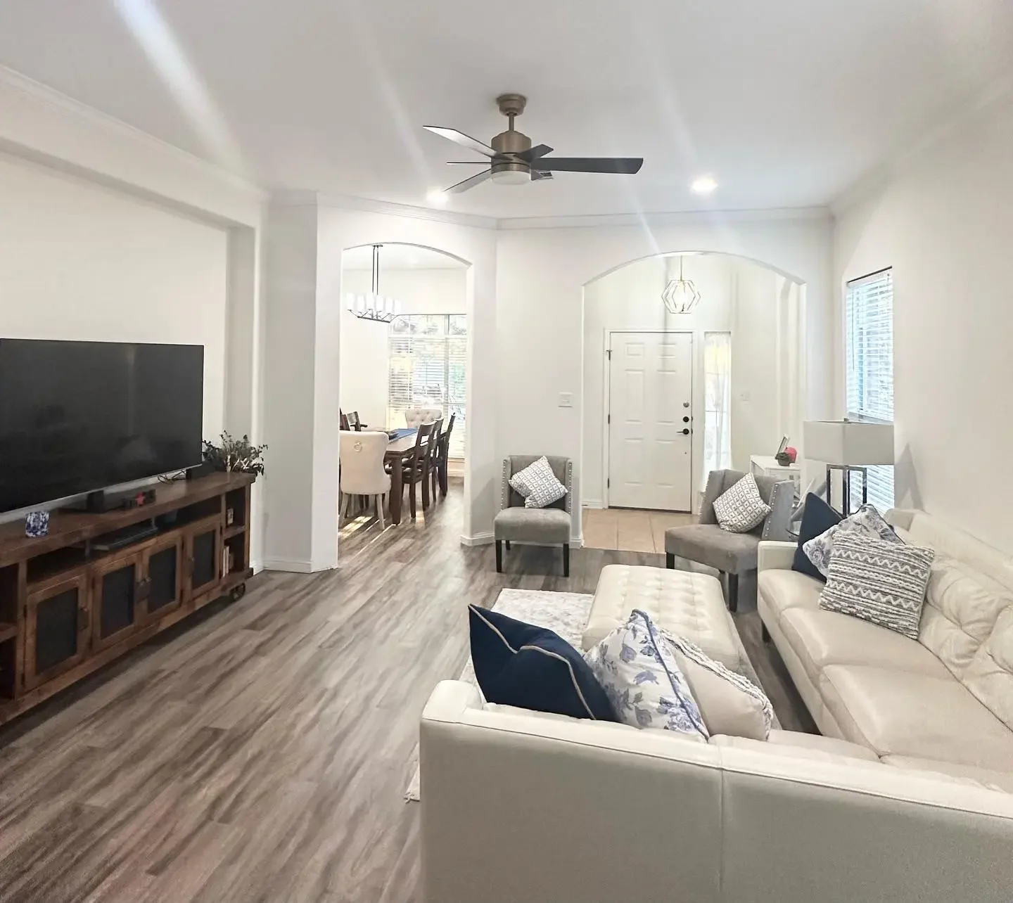Living room with white walls, hardwood floors, a ceiling fan, a large flat-screen TV on a wooden media console, a white sofa with decorative pillows, two gray armchairs, and a dining area with a wooden table and chairs visible through an archway.