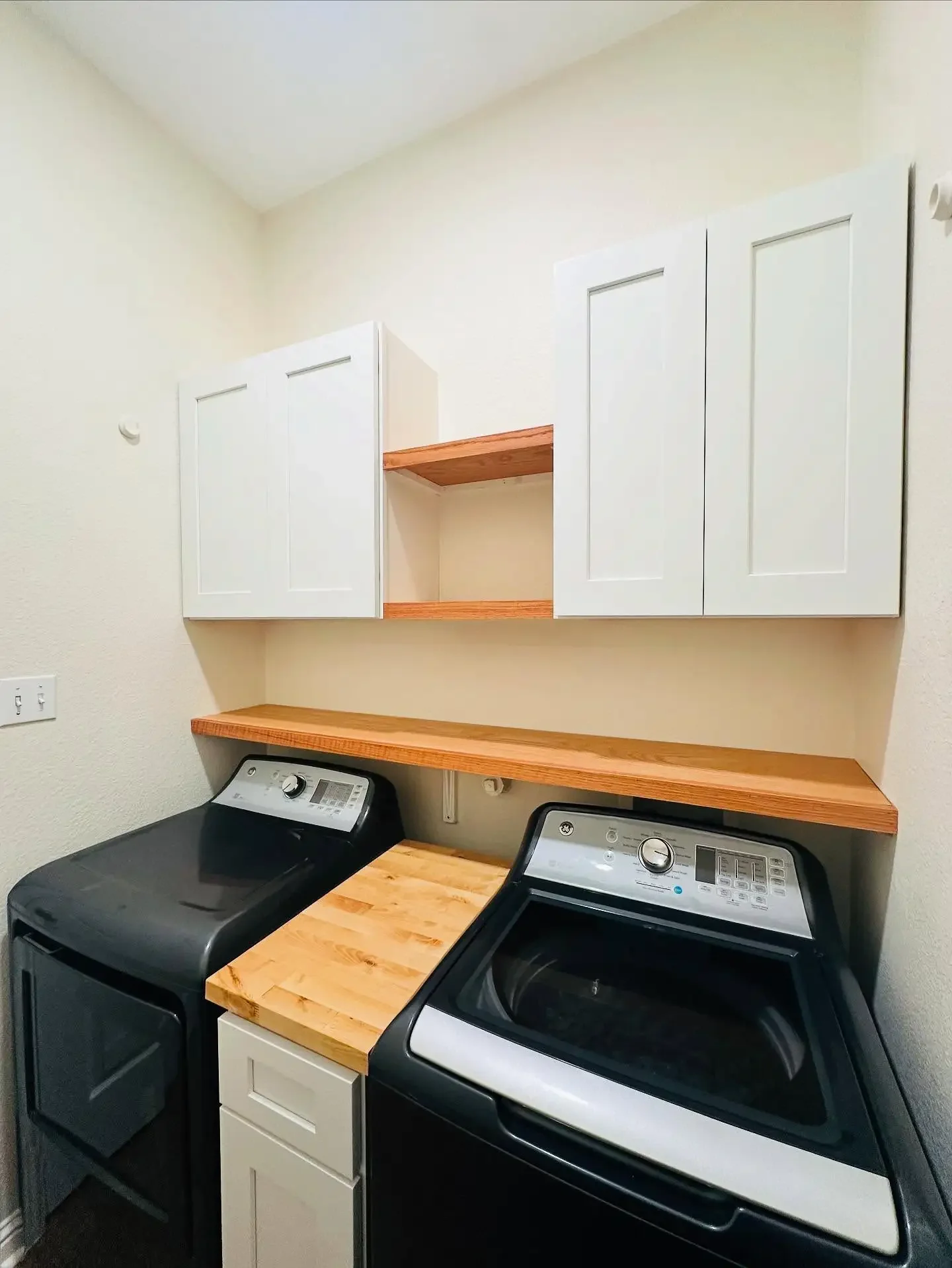 A laundry room with a black washing machine, a black dryer, a wooden countertop, and white wall cabinets.