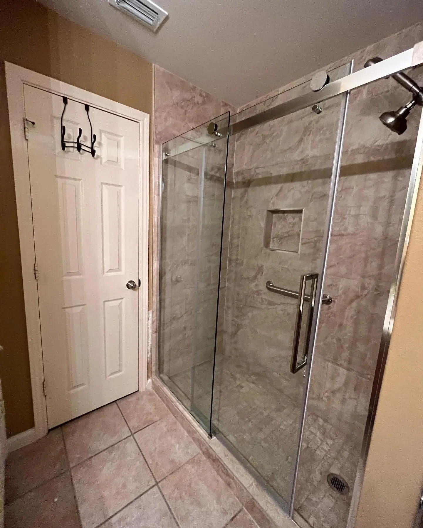 A bathroom with a glass shower enclosure, a chrome handle, a showerhead, and a built-in shower niche visible inside. The shower is tiled with beige marble tiles. Next to the shower, there is a white door and a coat hook rack on the wall.