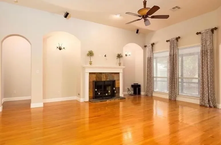 Living room with hardwood floors, a fireplace with a stone surround and white mantel, three large windows with patterned curtains, a ceiling fan, wall-mounted lights, and minimal decorations.