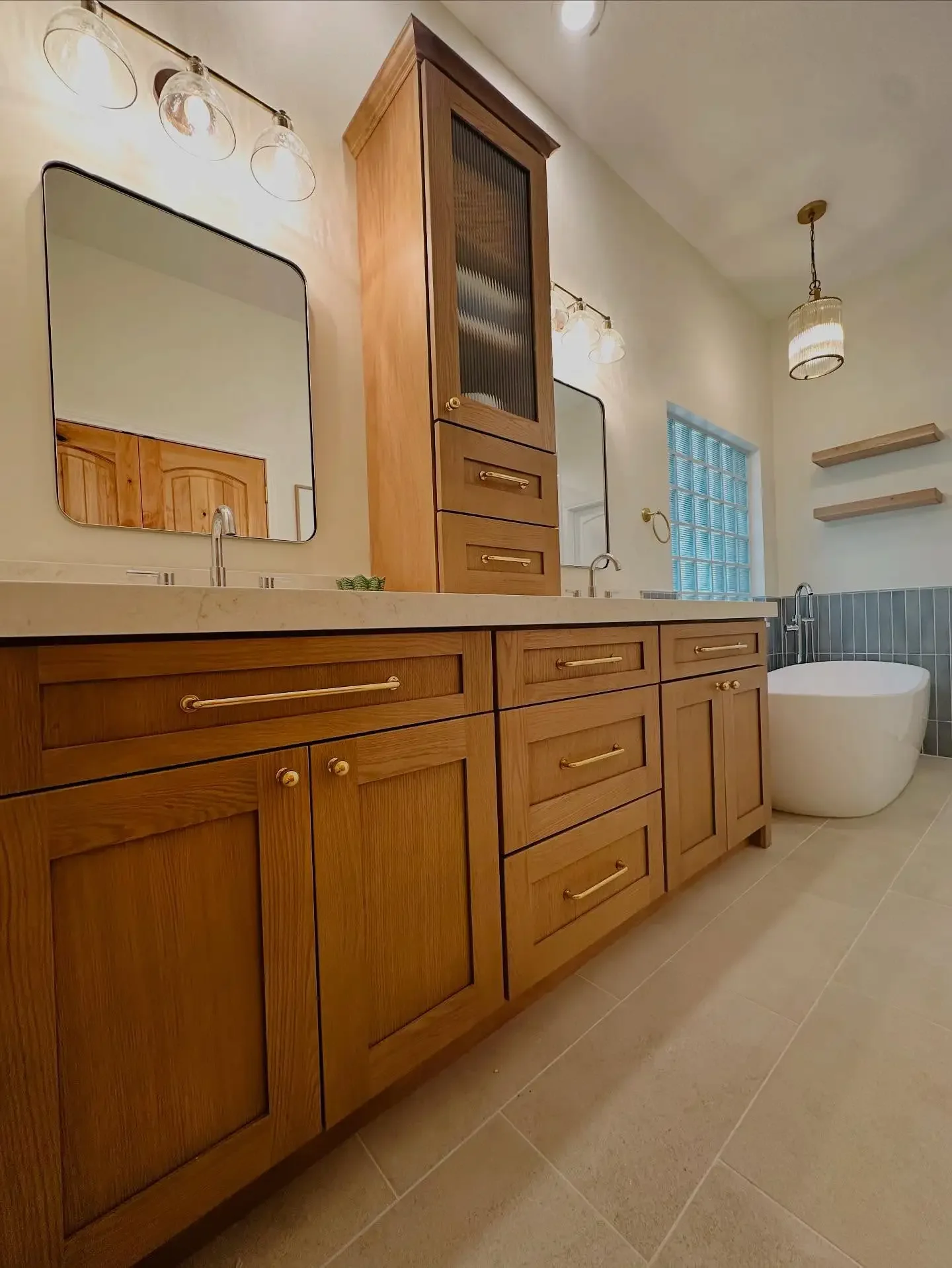 Bathroom with wooden cabinets, two mirrors, a freestanding bathtub, and wall-mounted shelves, illuminated by pendant and wall lights.