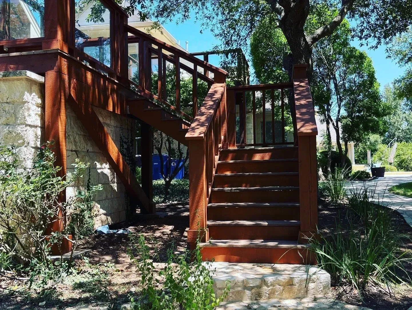 Wooden stairs leading to a deck with railing, attached to a stone wall, surrounded by green trees and plants under a clear blue sky.