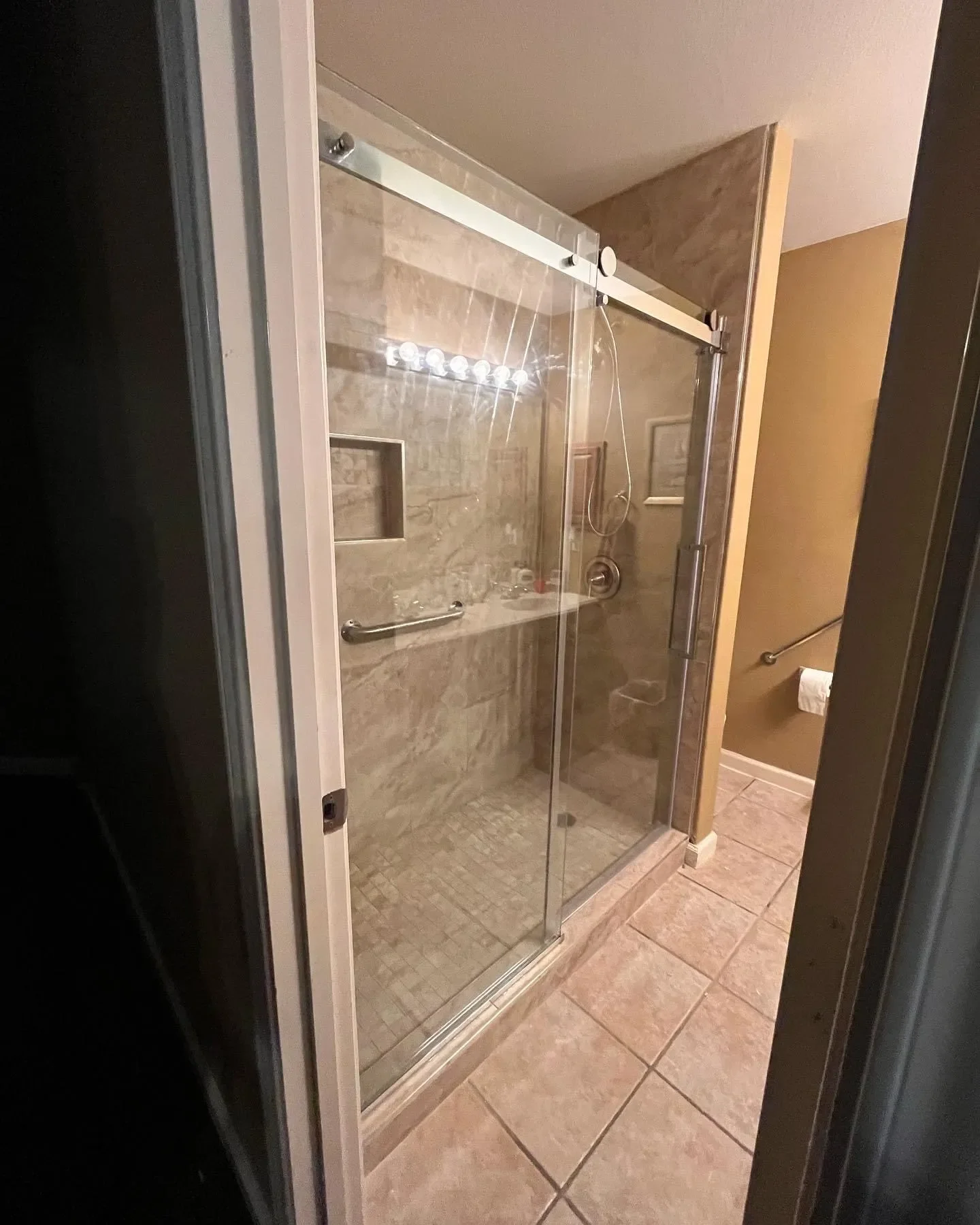 A bathroom with a glass shower door, beige tiled walls, and a beige tiled floor. There is a built-in shelf inside the shower and a showerhead. A towel is hanging on a rail outside the shower, and a roll of toilet paper is on a holder near the wall.
