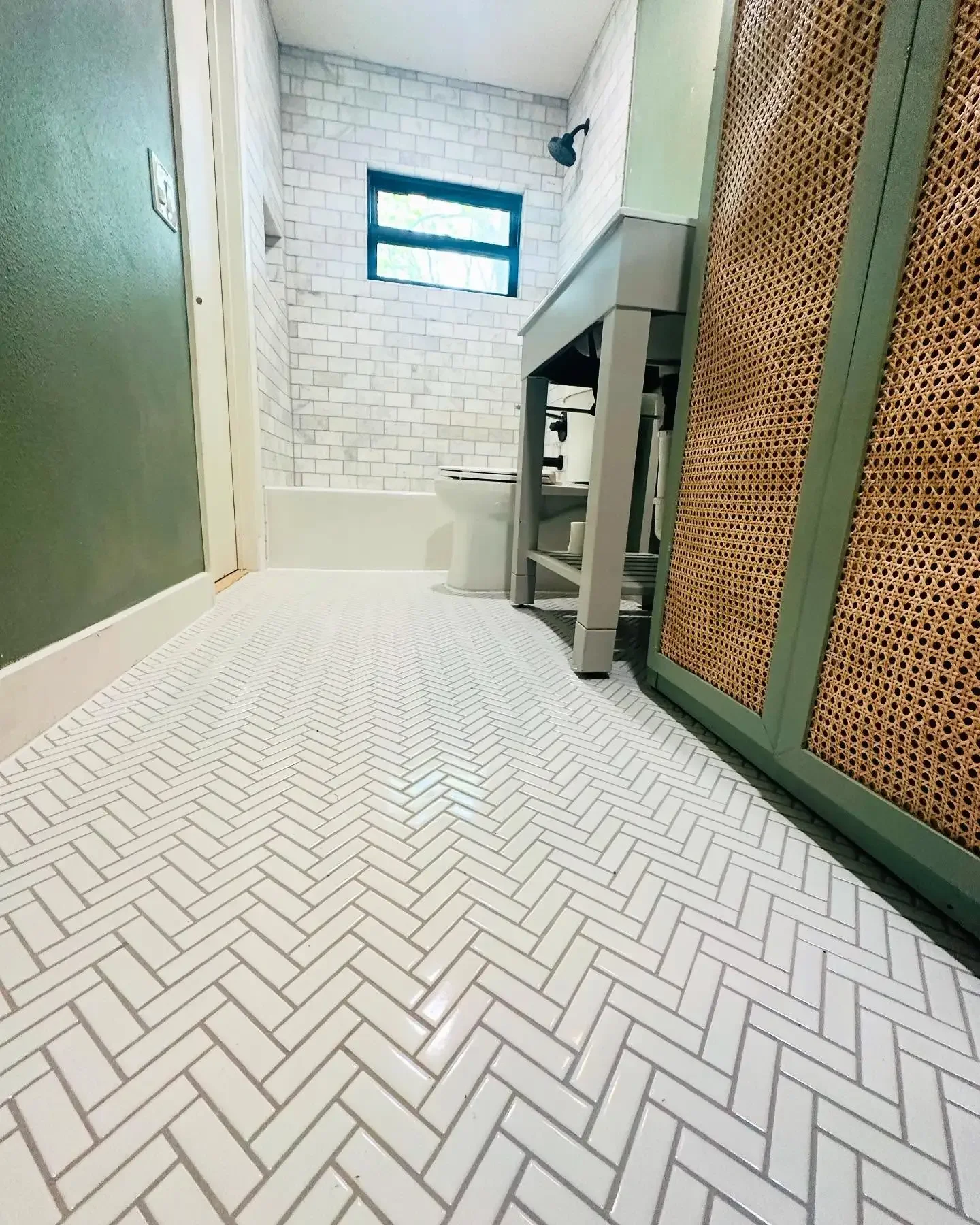Bathroom with white herringbone tile floor, green wall, small window, white brick shower wall, toilet, and vanity with a rattan panel cabinet.