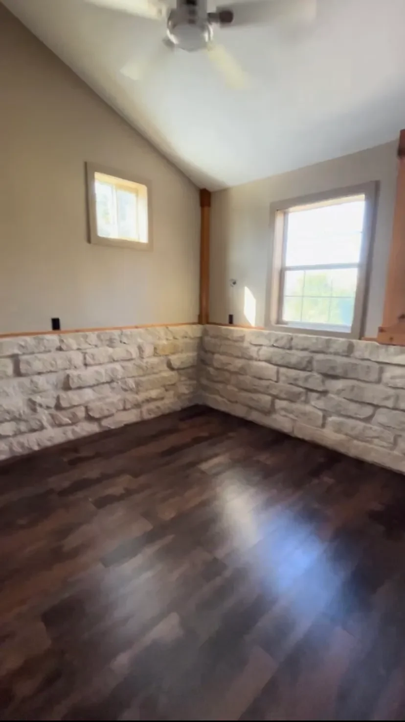 Interior of a room with a ceiling fan, two windows, a stone half-wall, and dark hardwood flooring.