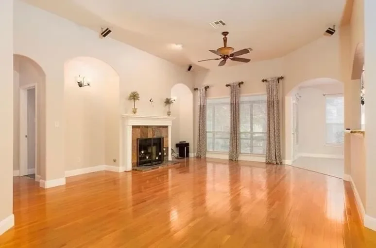 Empty living room with hardwood floors, a fireplace, windows with curtains, and a ceiling fan.