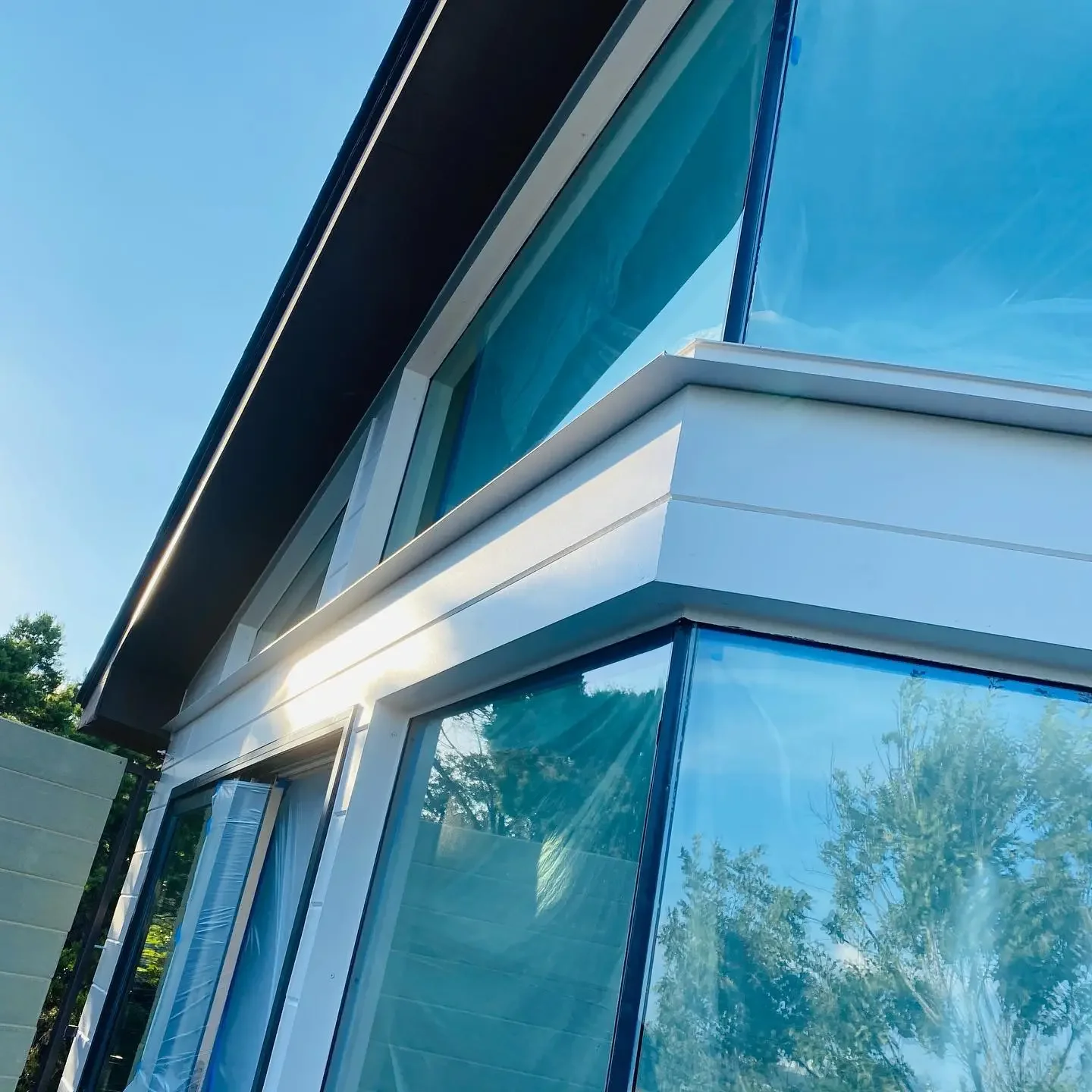 Close-up of a modern building with large glass windows and white structural elements, reflecting trees and the blue sky.