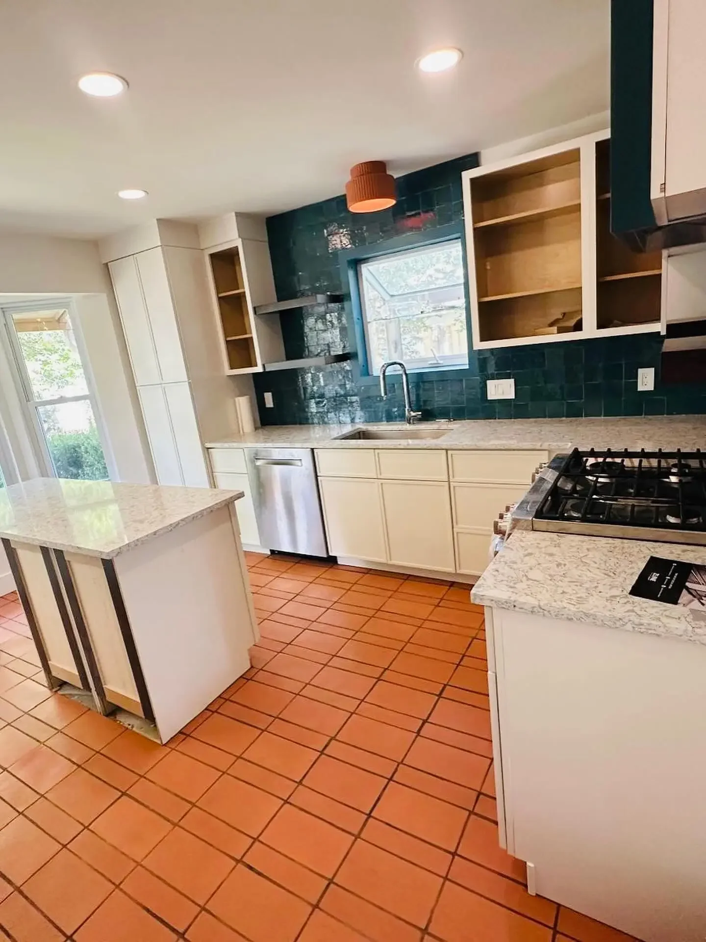 Kitchen with white cabinets, a granite countertop, a small island, and terracotta tile floors. There is a window above the sink, open shelving on the wall, and a stainless steel dishwasher. Recessed lighting is installed in the ceiling.
