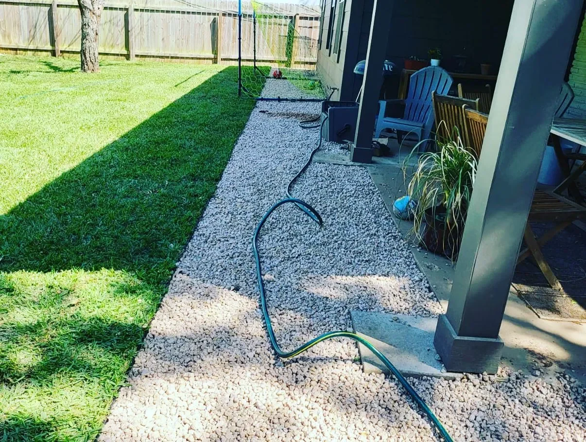 Backyard with a gravel pathway next to a grassy lawn, a fence, patio furniture, and gardening tools.