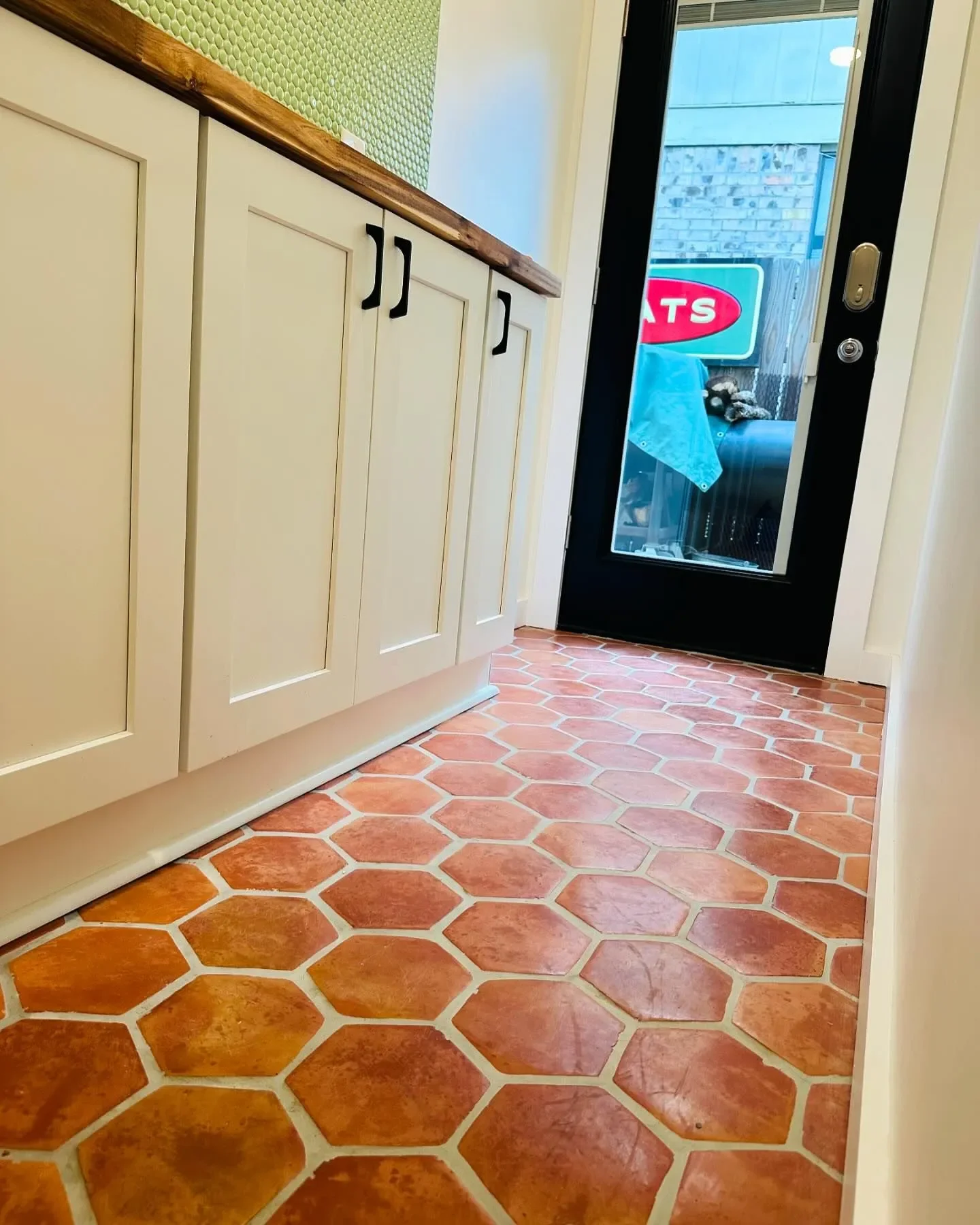 View of a kitchen with white cabinets, a wood countertop, terracotta hexagonal floor tiles, and a glass door leading outside, with a sign and some objects visible through the door window.