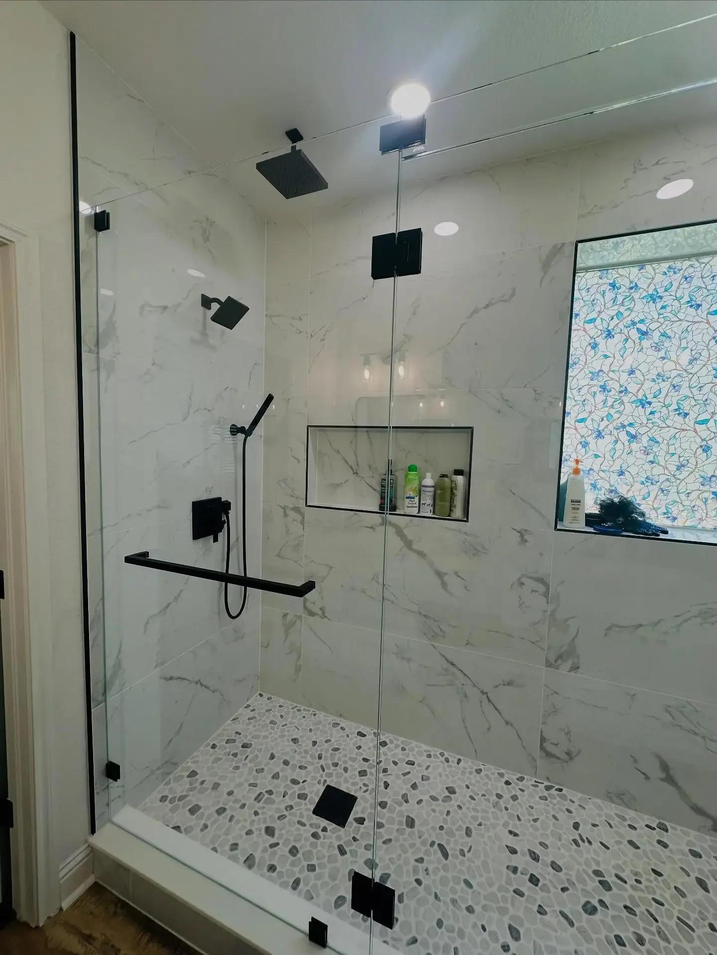 Modern walk-in shower with black fixtures, marble walls, pebble floor, and a glass door. Recessed shelf with toiletries and a small window with decorative patterned film.