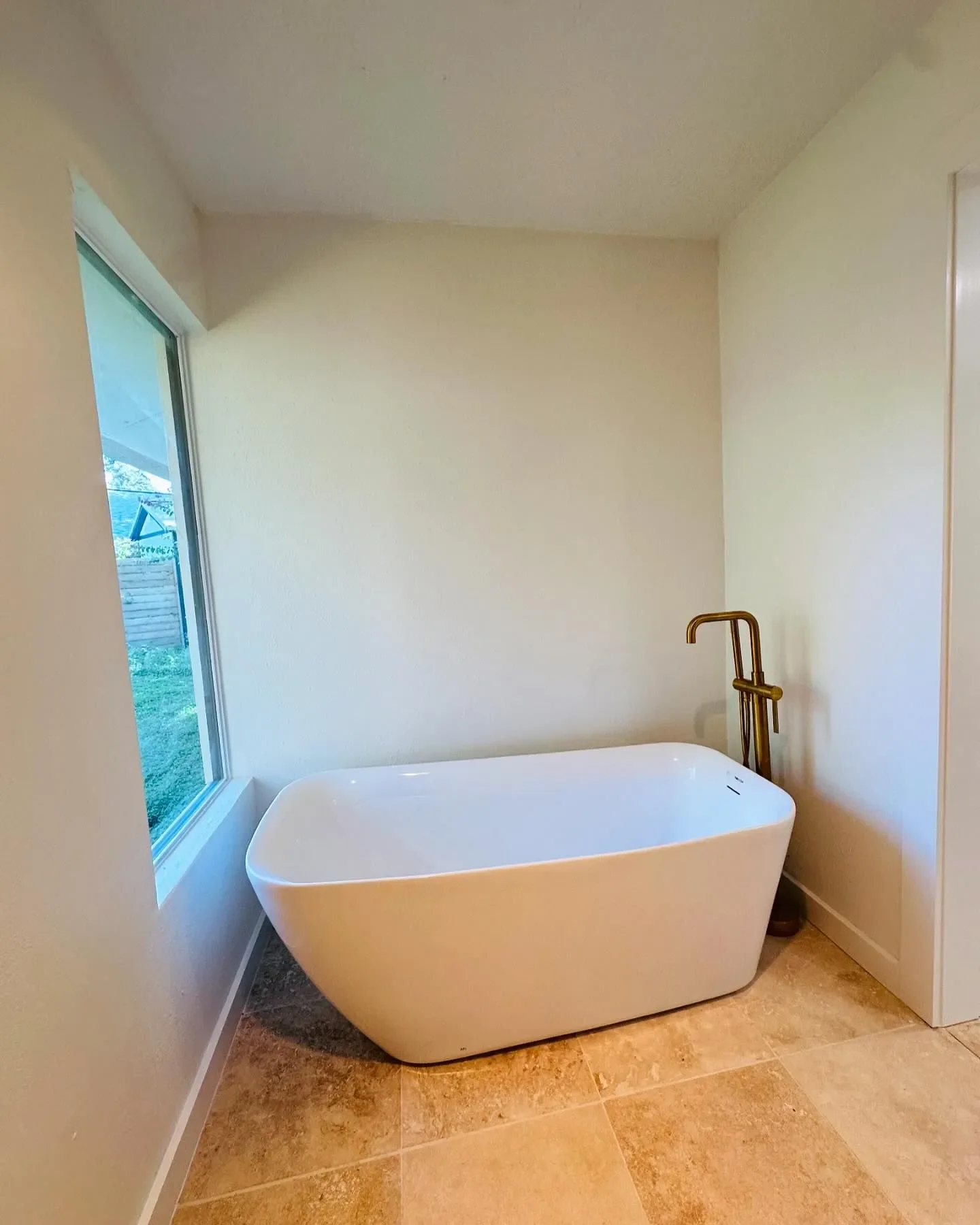 Modern white bathtub with floor-mounted gold faucet near a window in a bathroom with beige tiled floor and light-colored walls.