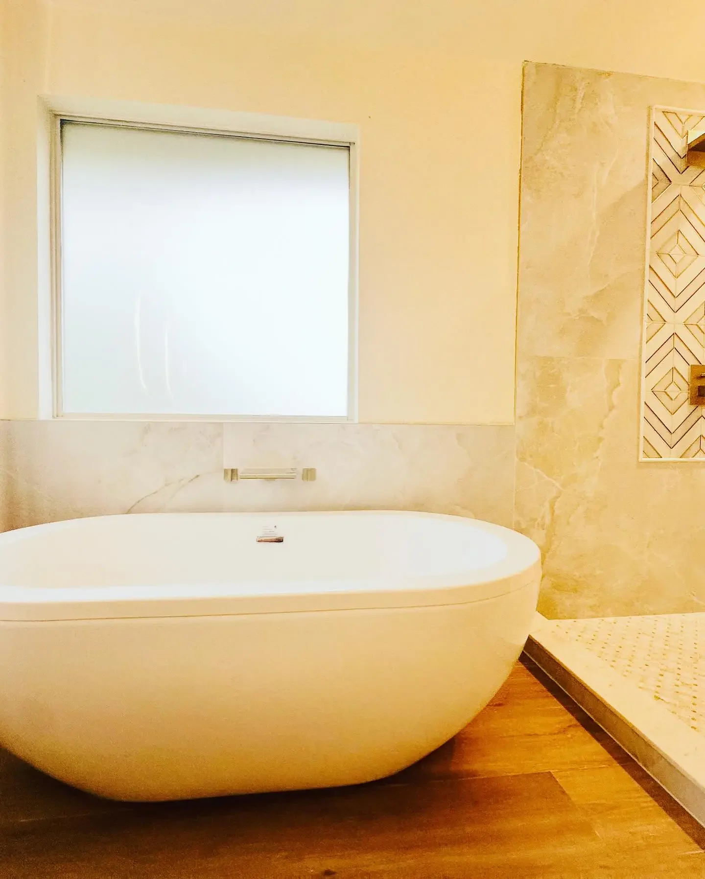 Modern bathroom with a freestanding bathtub, frosted window, marble wall, and wood-look flooring.