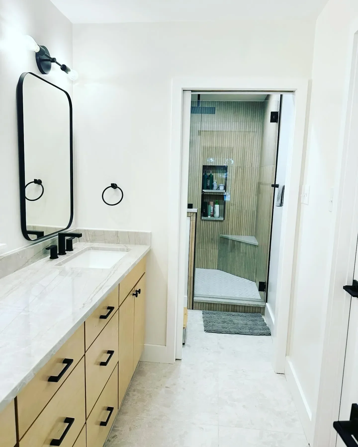 Bathroom with light wooden cabinet, black fixtures, a mirror, a soap dispenser, and a sliding glass shower door with wood paneling inside.
