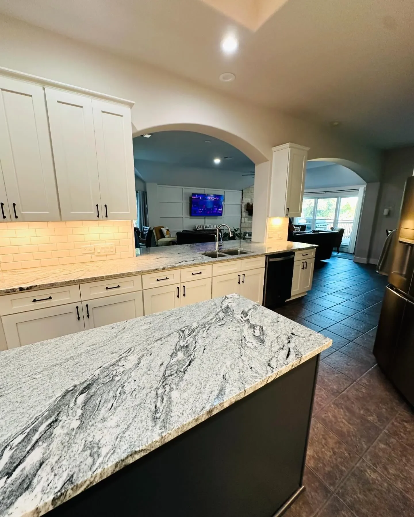Modern kitchen with white cabinetry, granite countertops, and dark tiled flooring, opening into a living room with a TV and seating area.