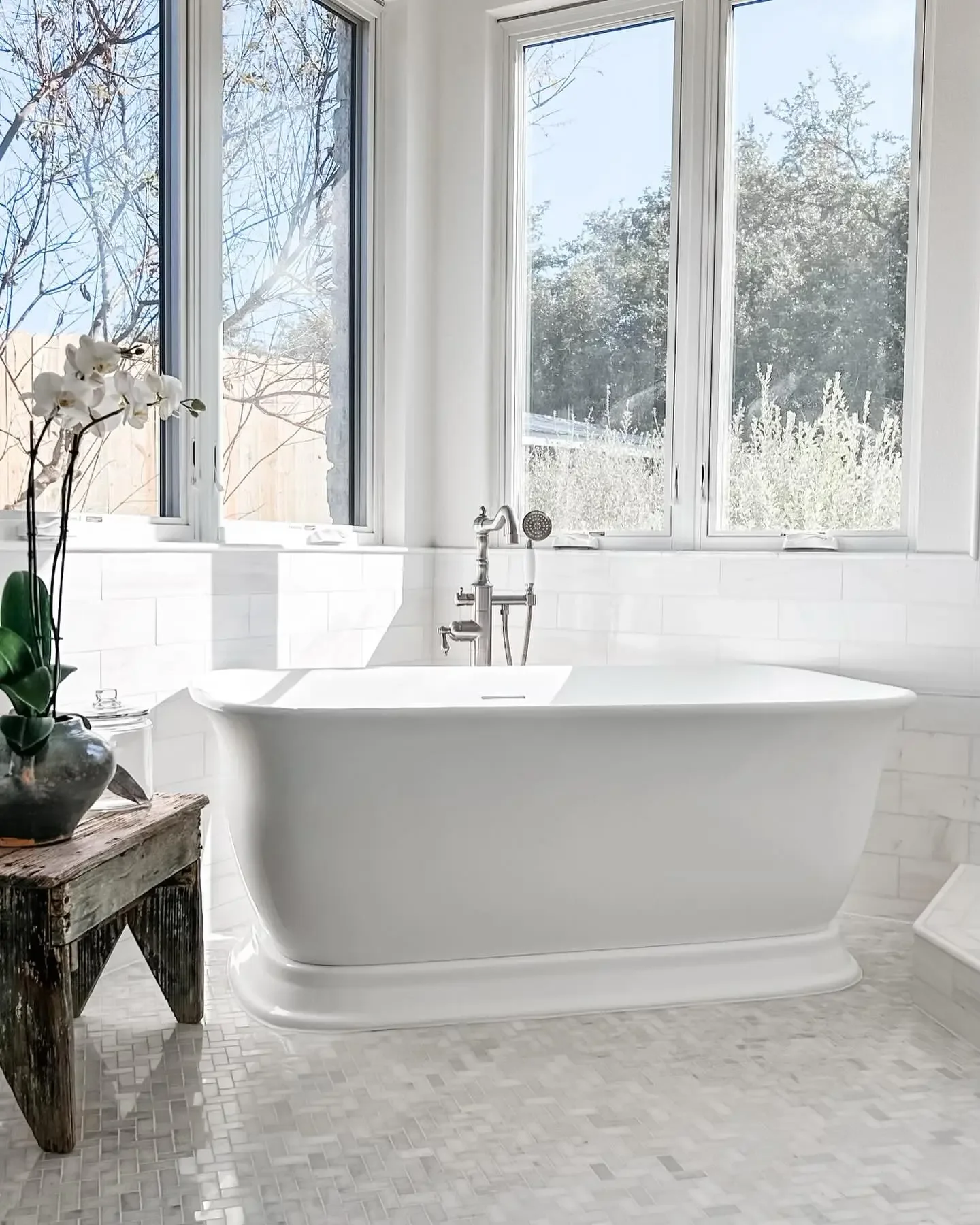 A white freestanding bathtub in a bright bathroom with large windows, a wooden side table with a potted orchid, and a tiled floor.