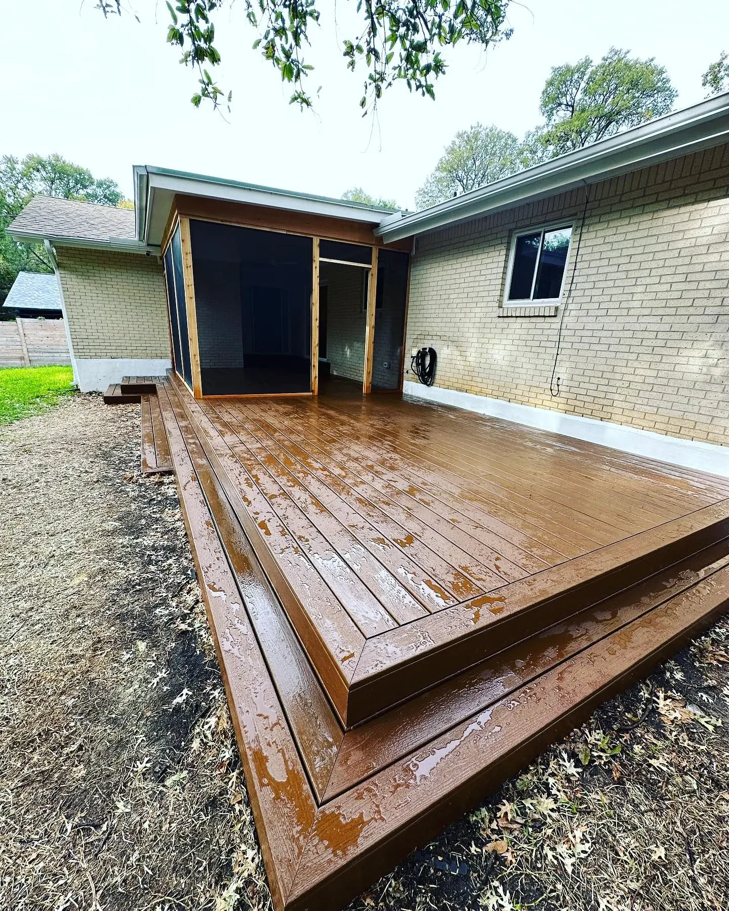 Freshly installed brown wooden deck attached to a brick house with a screened porch, wet from rain, with greenery in the background.