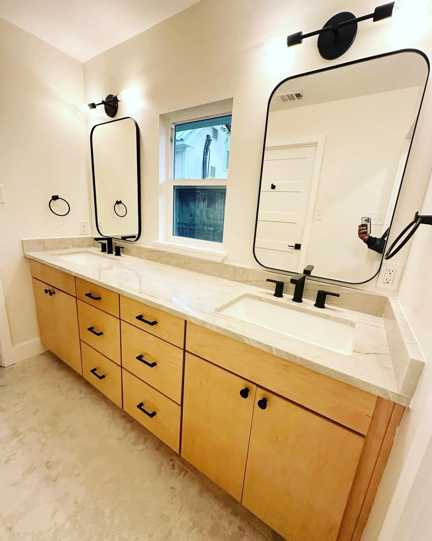 Modern bathroom vanity with dual sinks, black fixtures, light wood cabinets, two large rectangular mirrors, and a window in between, with beige marble countertop and beige floor.