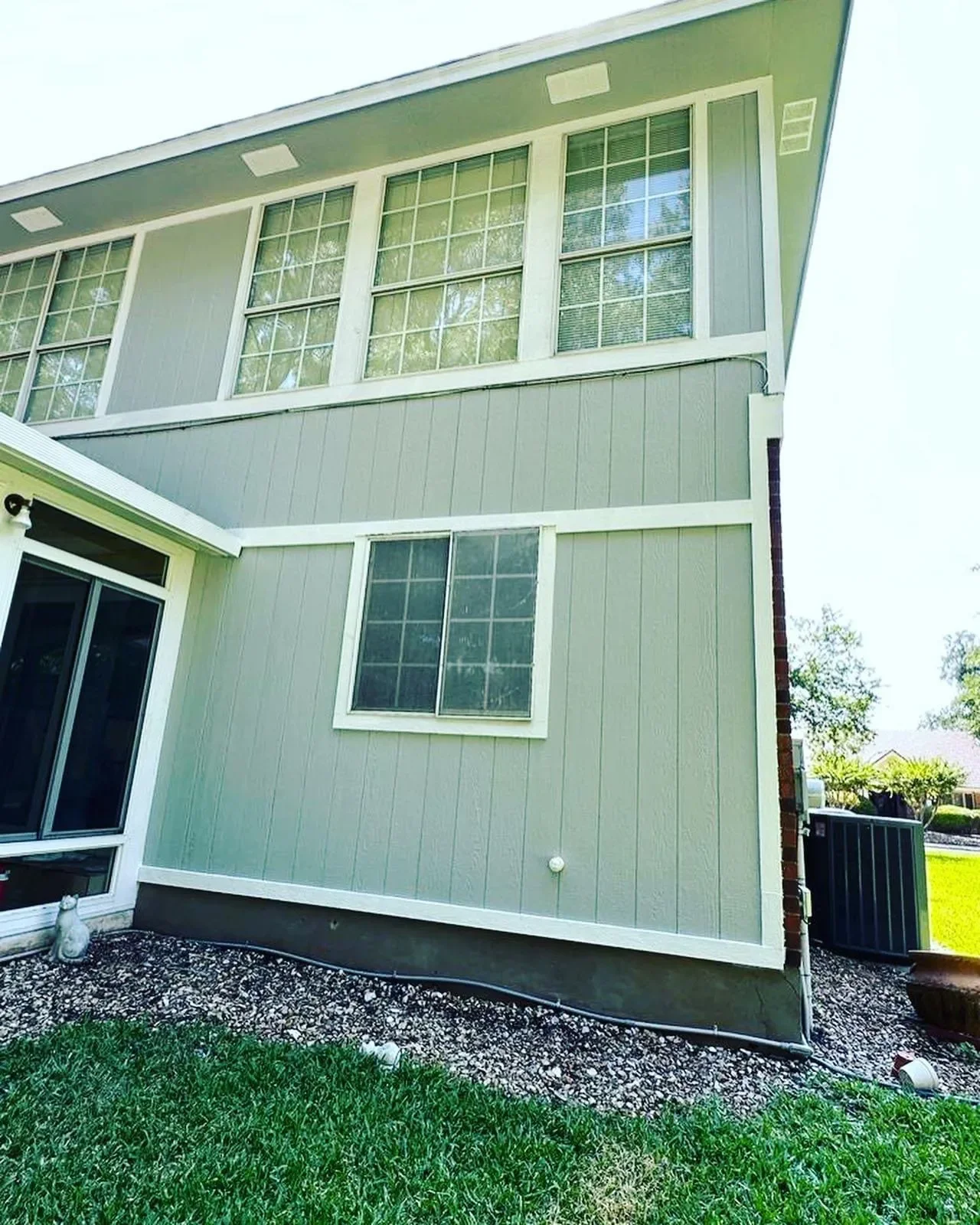 The back side of a two-story house with gray siding, multiple windows, a sliding door, and a small grass area with gravel near the foundation.