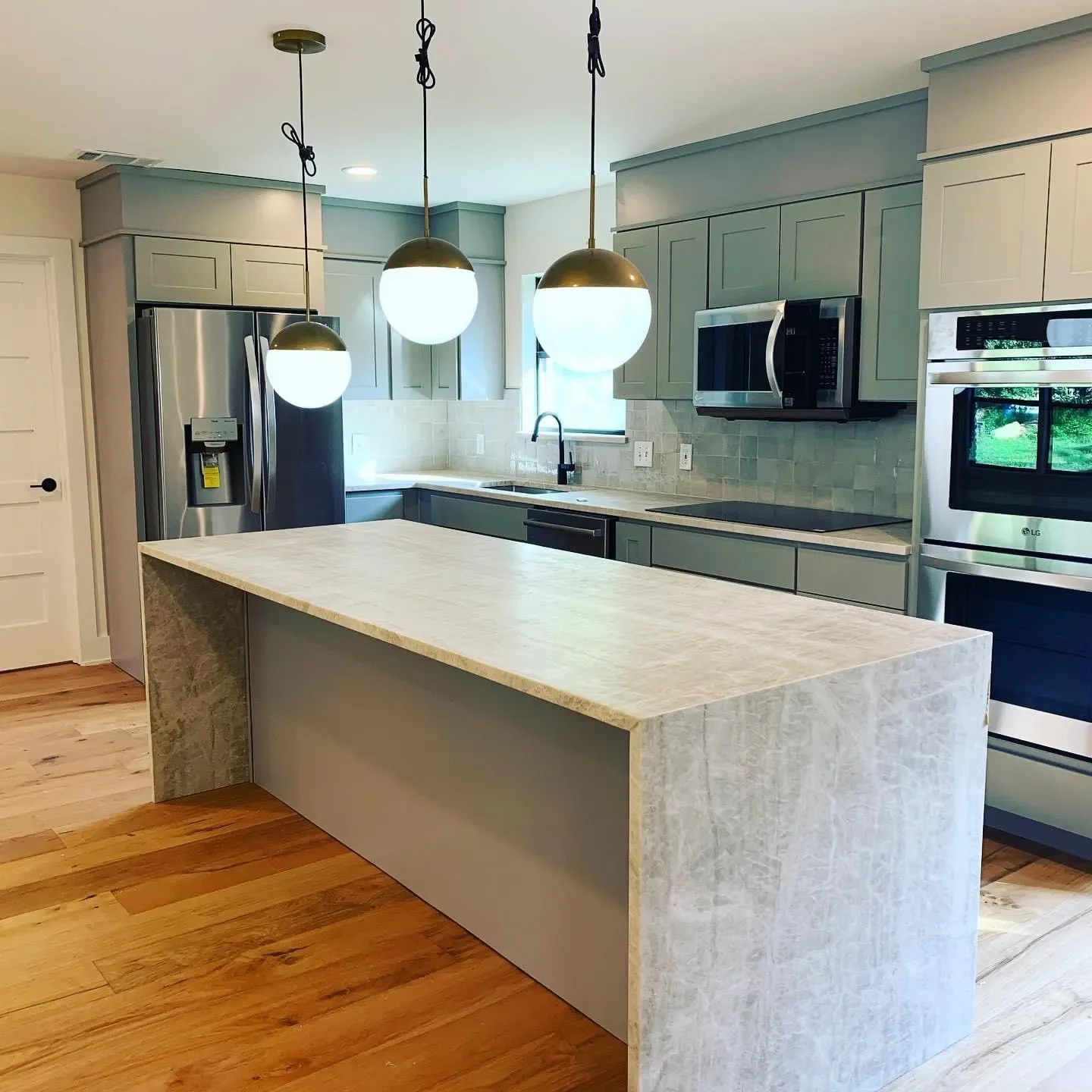 Modern kitchen with light green cabinets, stainless steel appliances, a large island with a light-colored countertop, and three hanging pendant lights with round white and brass fixtures.