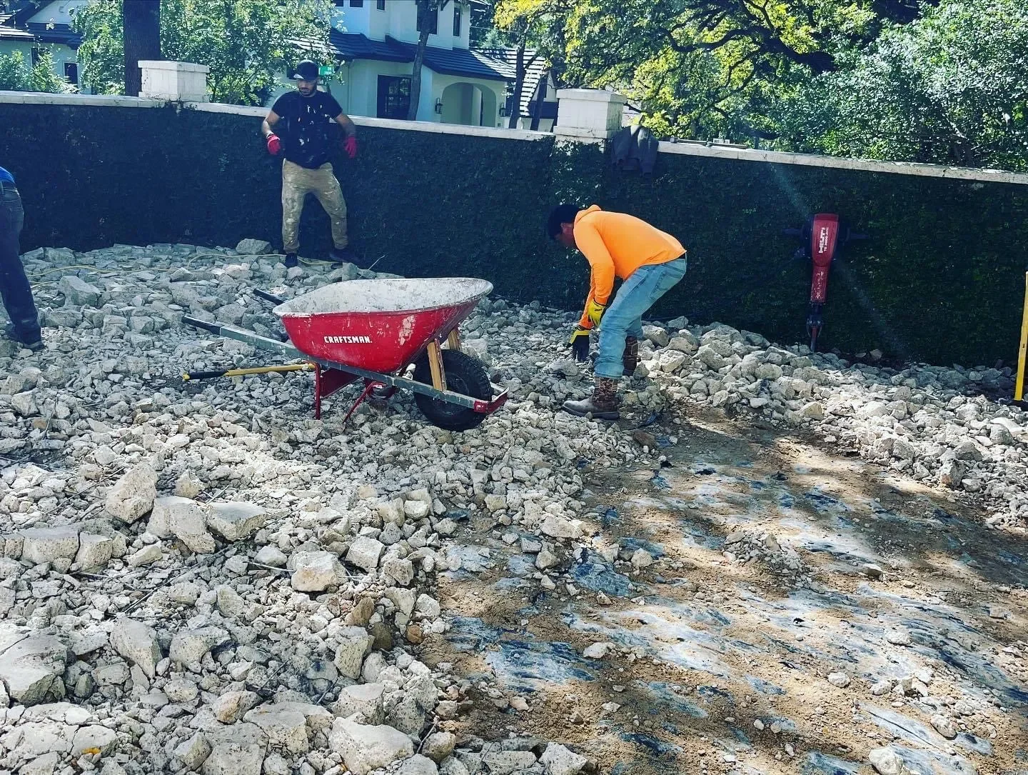 Two construction workers breaking rocks on a construction site with a wheelbarrow and tools, near a fence with trees and houses in the background.