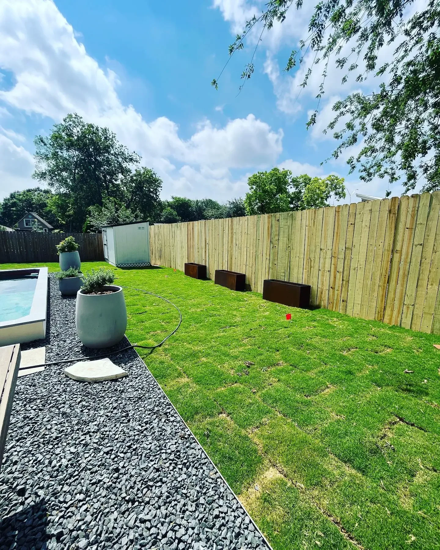 Backyard with grass, wooden fence, potted plants, shed, and a pool on the left under a partly cloudy sky.