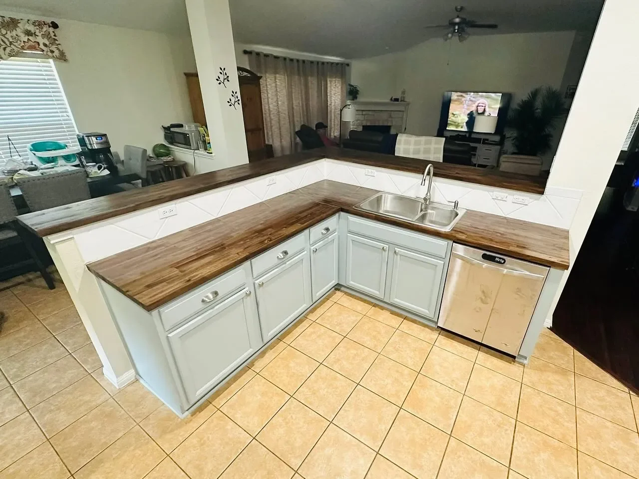 Kitchen with light blue cabinets, wooden countertops, a stainless steel sink, and a small dishwasher, with a view into living and dining areas.