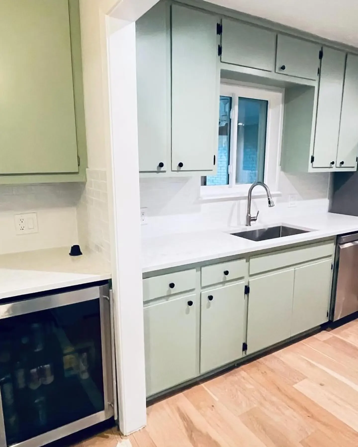 A kitchen with light green cabinets, white countertops, and a stainless steel sink with a window above it. There is a small wine cooler or beverage fridge on the left and wooden flooring.