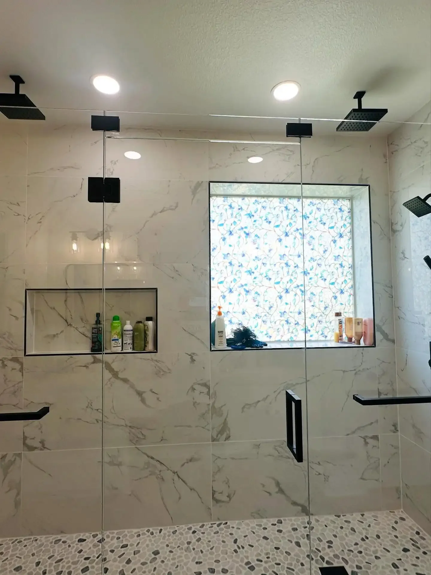 A modern walk-in shower with marble walls, built-in shelves containing toiletries, a window with a decorative patterned cover, and black fixtures including shower heads and handles.