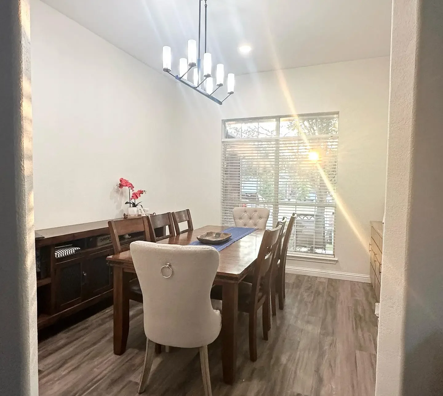 Dining room with a wooden table, six chairs, a decorative centerpiece, window with blinds, and sunlight streaming in.