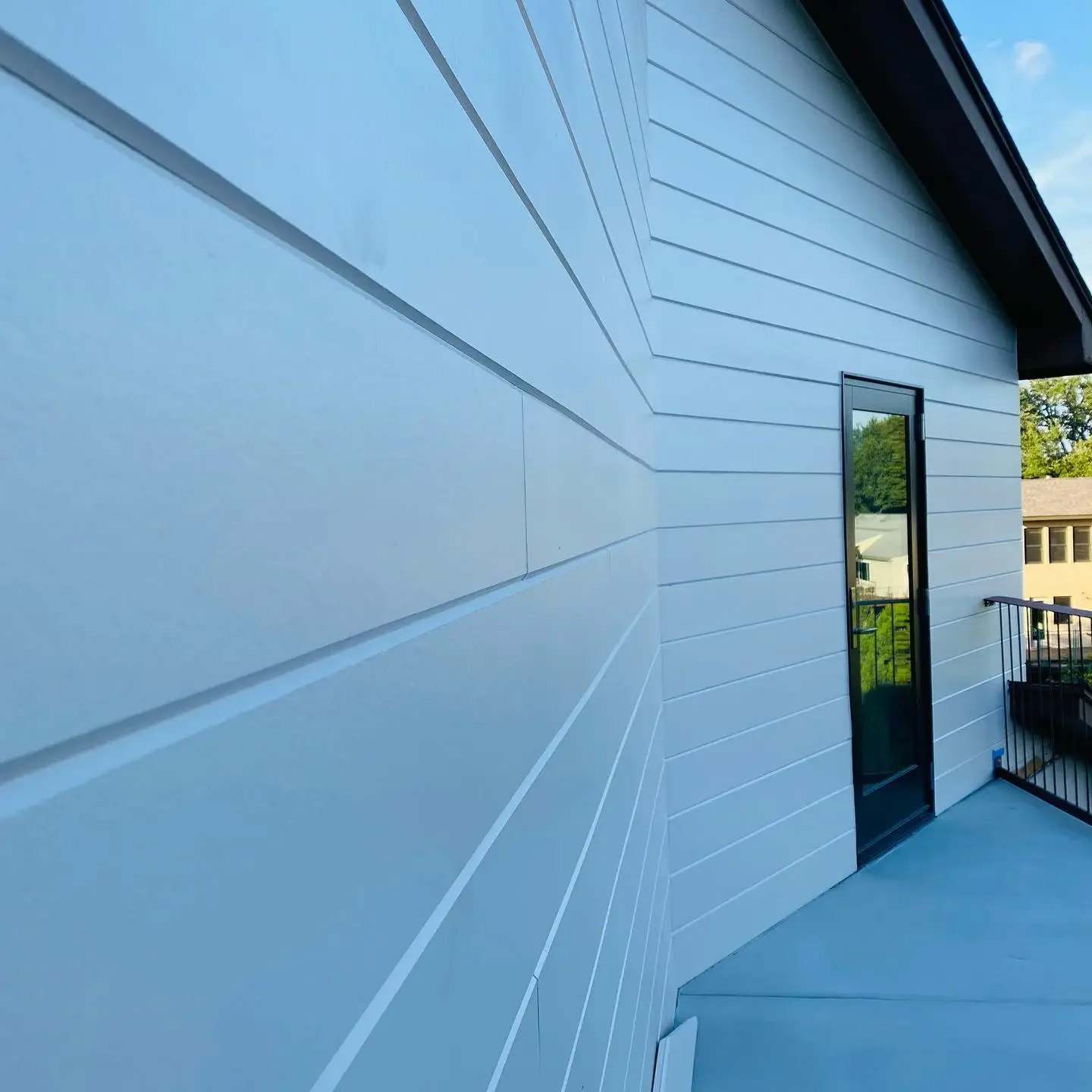 Exterior side of a modern house with blue siding, a door with large glass pane, a small balcony with black railing, and neighboring houses in the background under a partly cloudy sky.