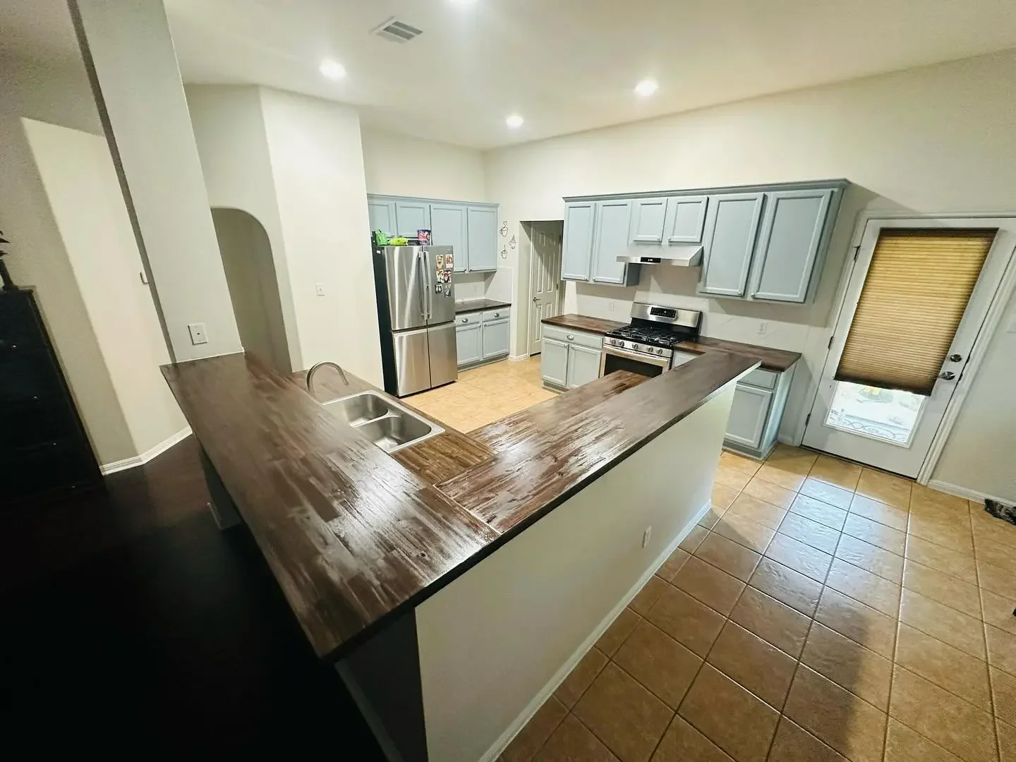 A kitchen with light blue cabinets, stainless steel refrigerator, gas stove, wooden countertops, a sink, and a door with a window and a beige blind, tiled floor, and recessed ceiling lights.