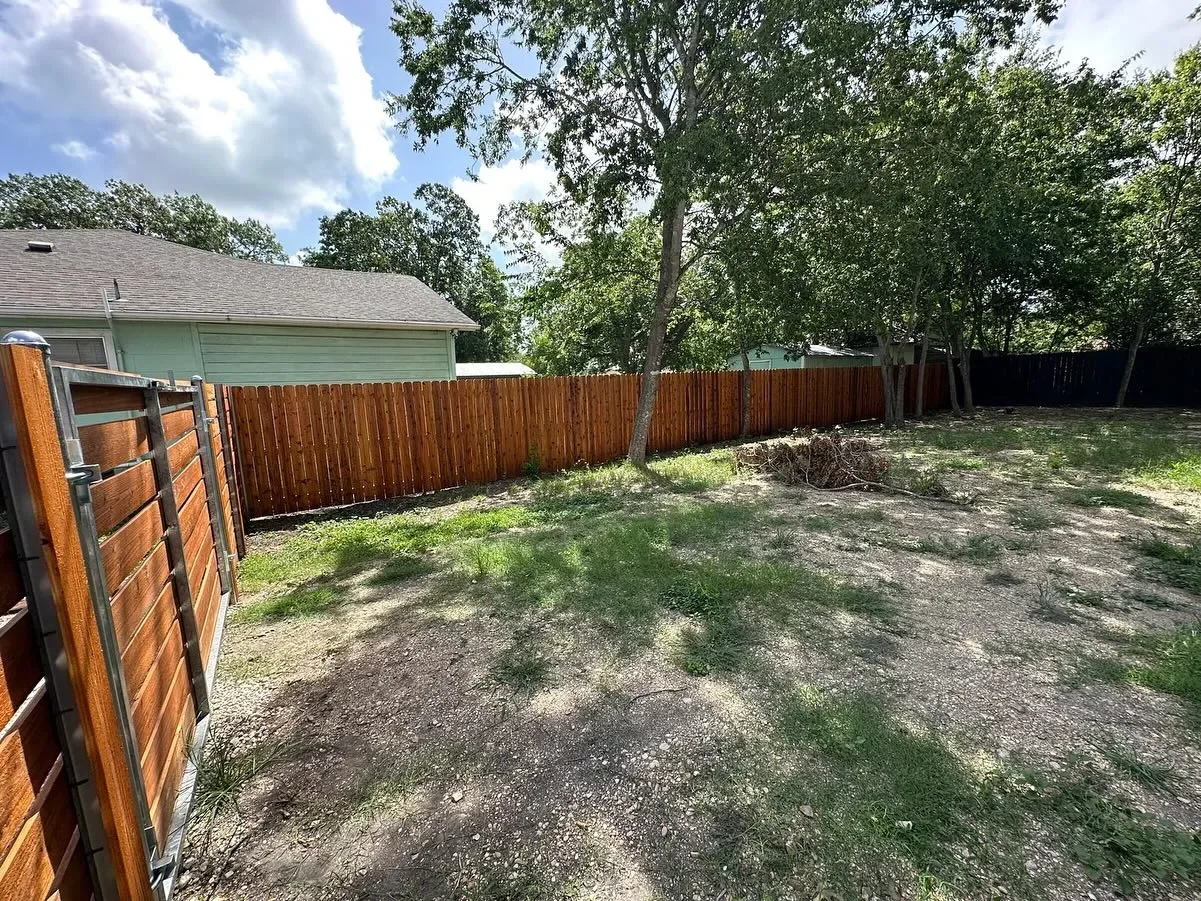 Backyard with a new wooden fence, trees, and patches of grass and dirt under partly cloudy sky.