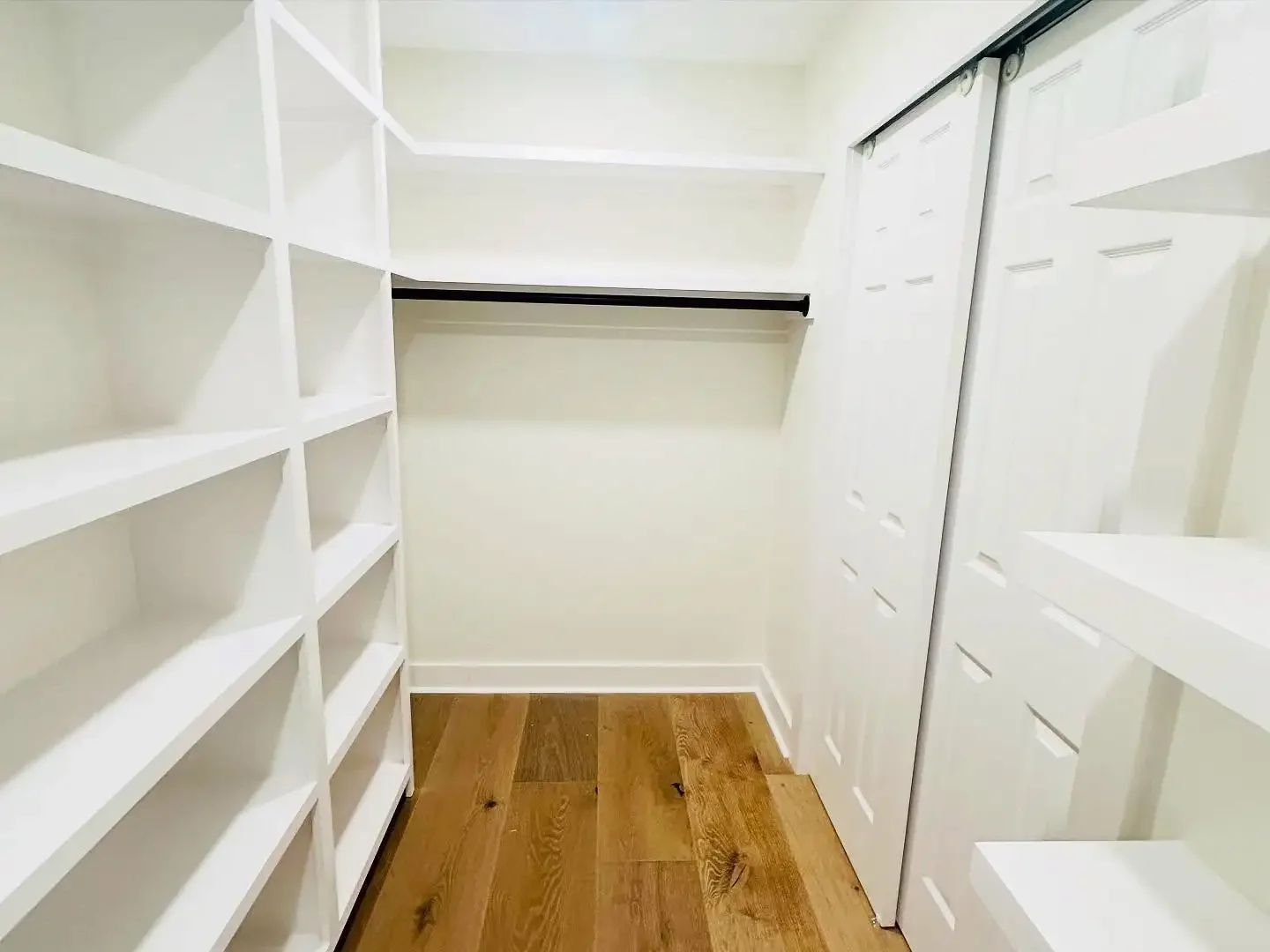 Empty walk-in closet with white built-in shelves on the left side, a hanging rod on the back wall, white sliding door cabinets on the right side, and a hardwood floor.