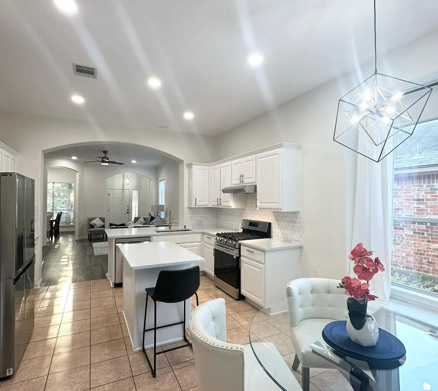 Open-concept kitchen with white cabinets, stainless steel appliances, and a kitchen island with a black chair. Adjacent dining area features a round glass table with white chairs and a potted pink flower. Bright lighting and natural light through a w