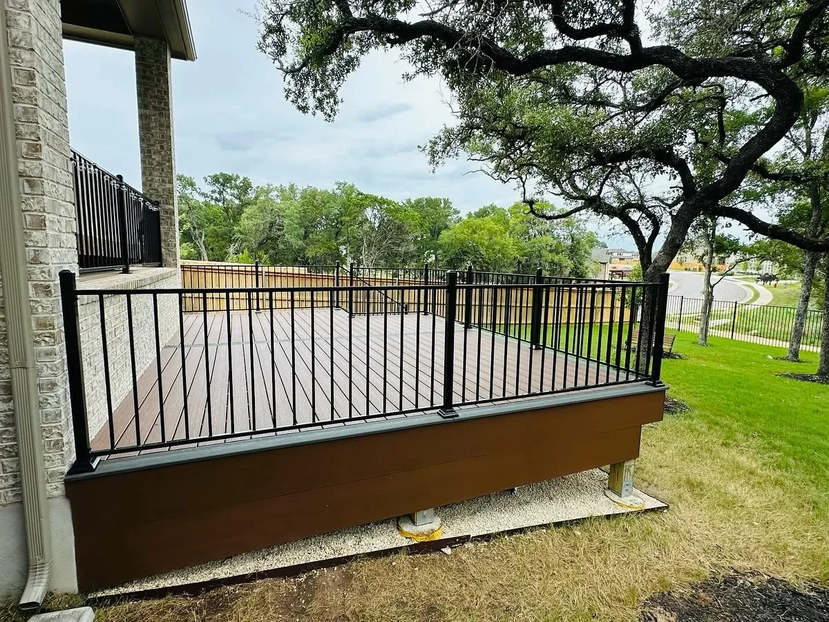 Newly built wooden deck with black metal railing attached to a brick house, overlooking a grassy yard with trees and a street.