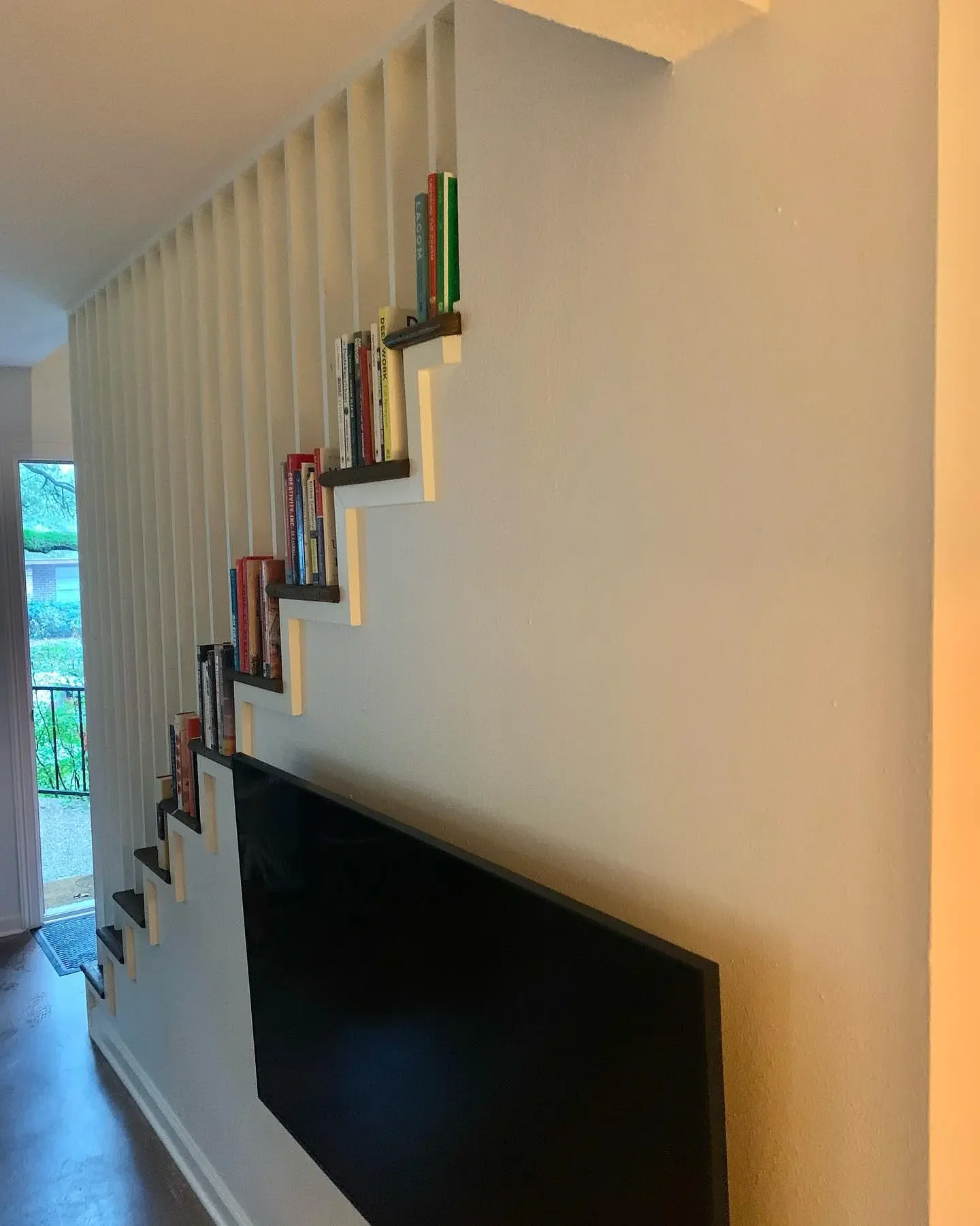 Wall with staggered black and white bookshelf containing books, a large flat-screen television below, next to a window with outdoor view.