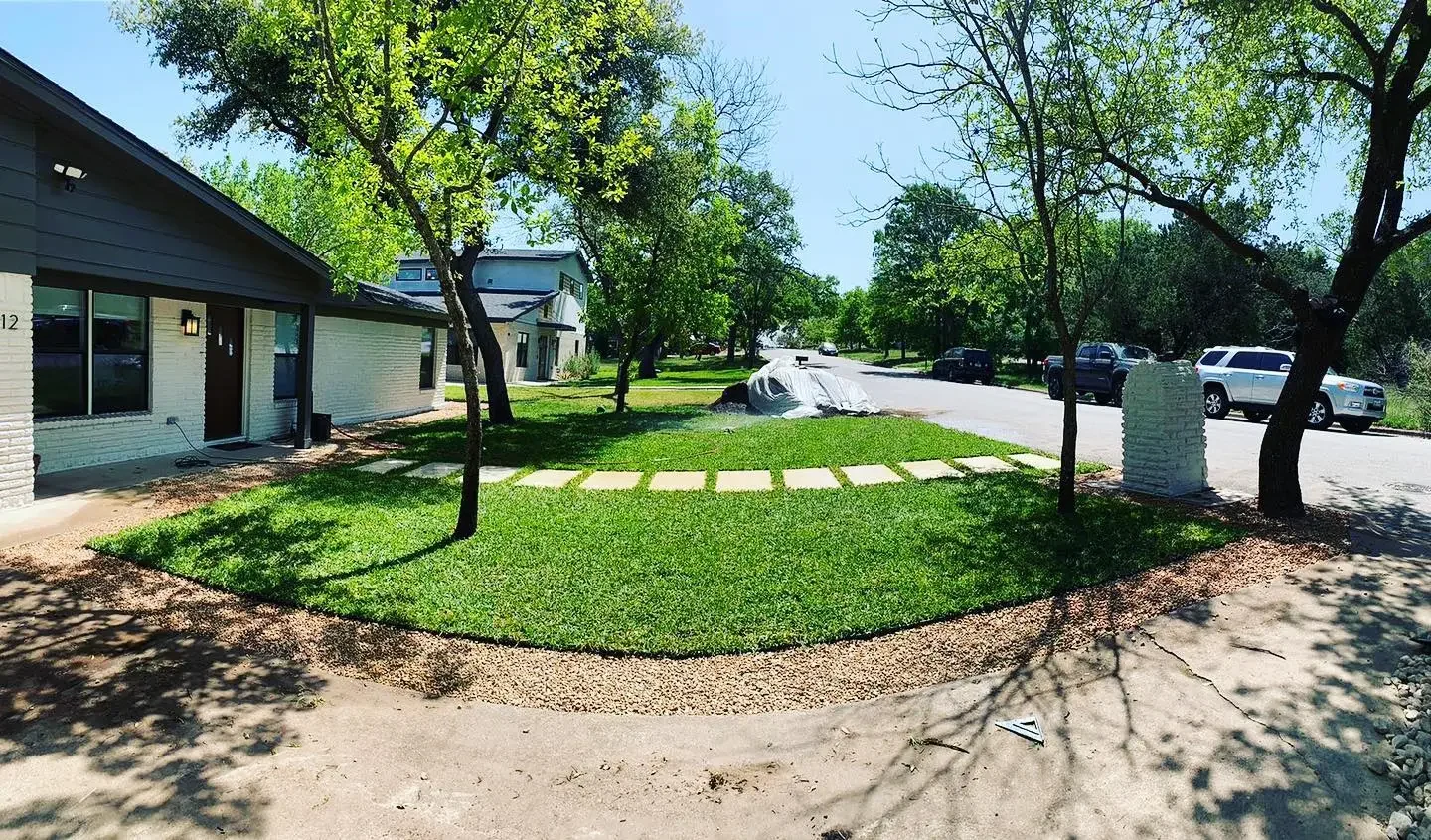 A front yard with a grassy lawn and stepping stones leading to the front of a house. Trees shade part of the yard and street, with parked cars visible along the curb.