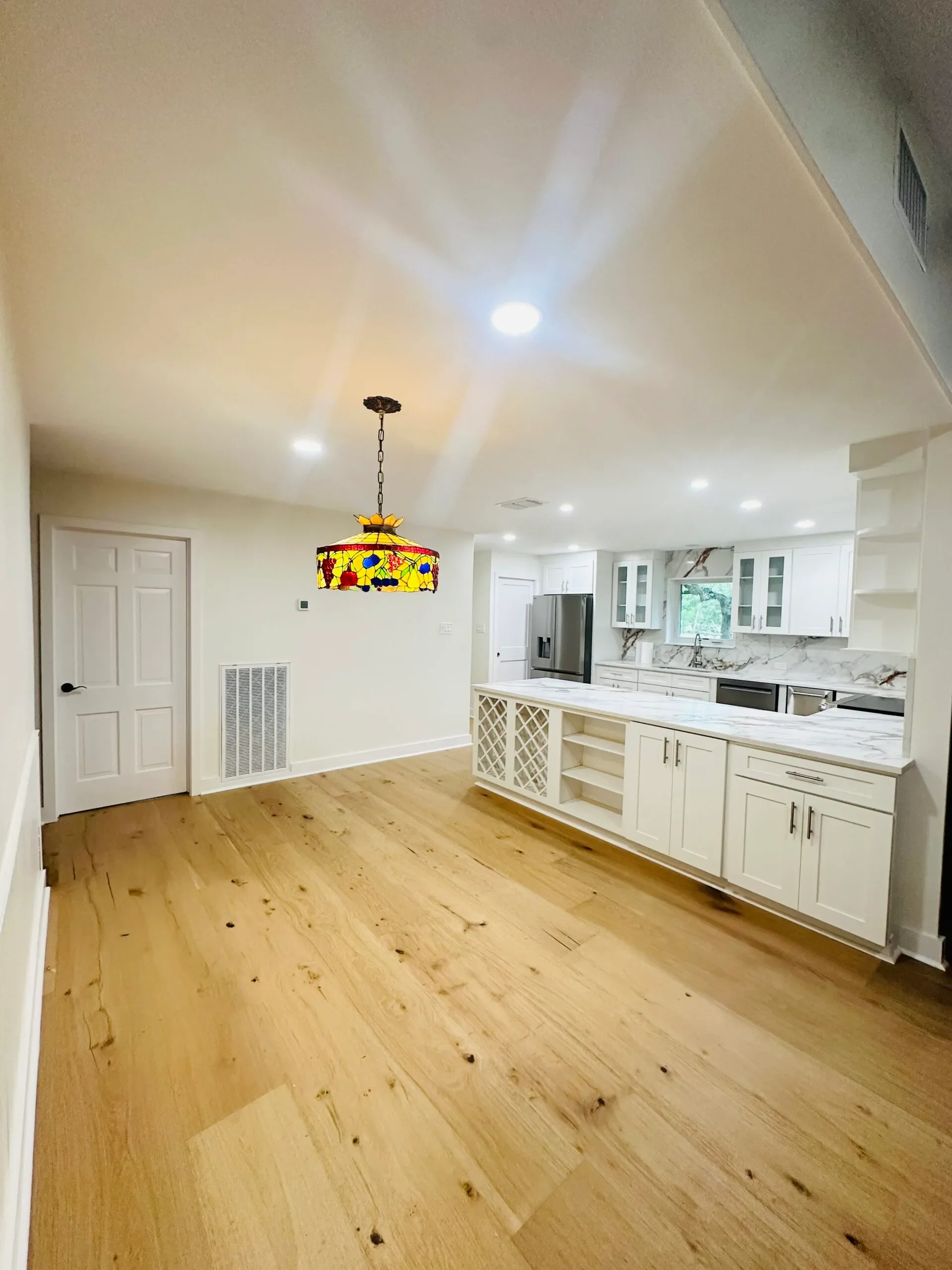 Bright, open kitchen with white cabinets, marble countertops, stainless steel appliances, a window above the sink, hardwood floors, and a Tiffany-style stained glass hanging light fixture.