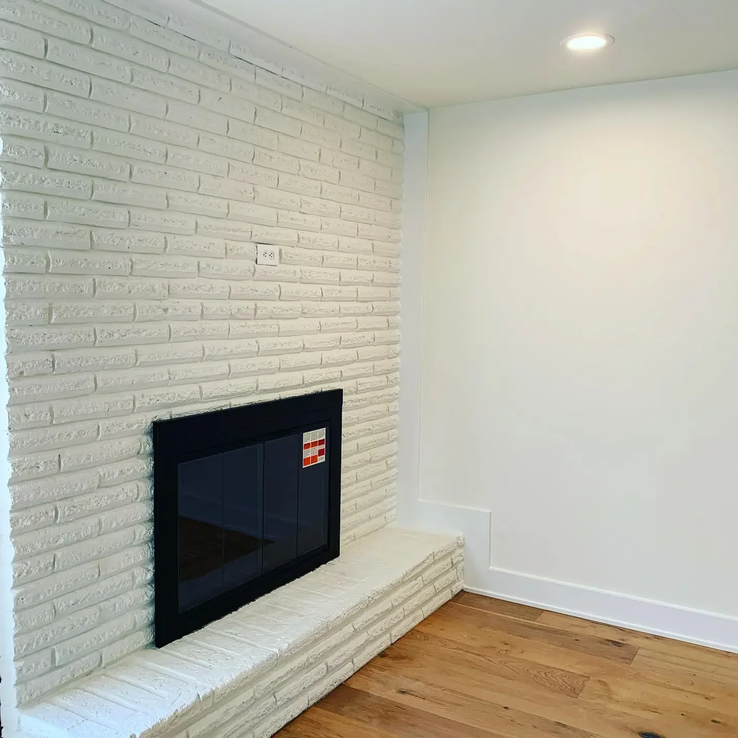 A white brick fireplace on a wall with a small red and white decorative tile, wooden floor, and white wall with a recessed ceiling light.