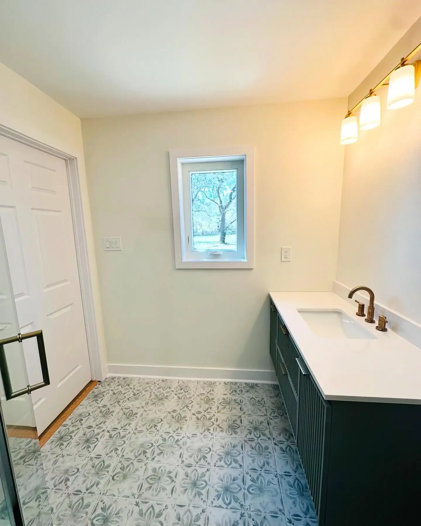 Empty bathroom with a small window, white walls, a dark vanity with a white countertop and sink, a brass faucet, and patterned tile flooring.
