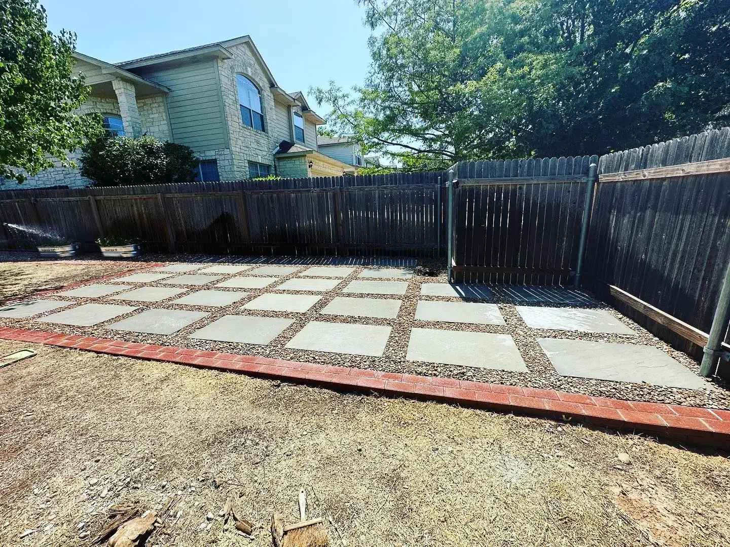 Backyard patio with large concrete tiles arranged in a grid, bordered by red bricks, and surrounded by a wooden privacy fence. There are two raised garden beds with plants and a hose spray on the left. A house with white stone and siding is visible i